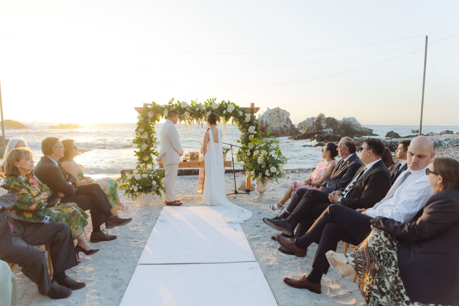 Elegant beach wedding ceremony with white aisle, floral arch, and seated guests at sunset.