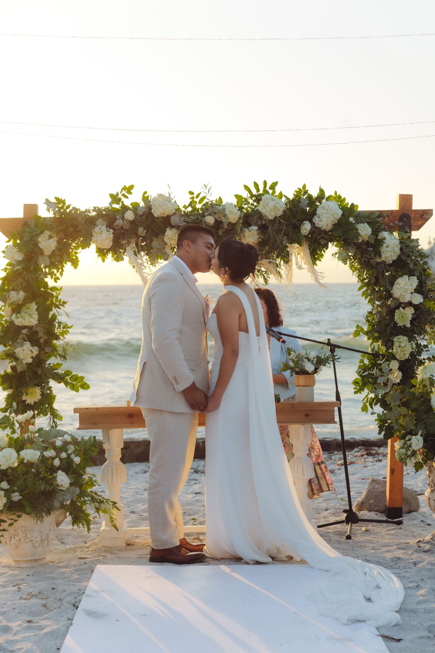 Bride and groom share first kiss at beach wedding under lush white floral arch at sunset.