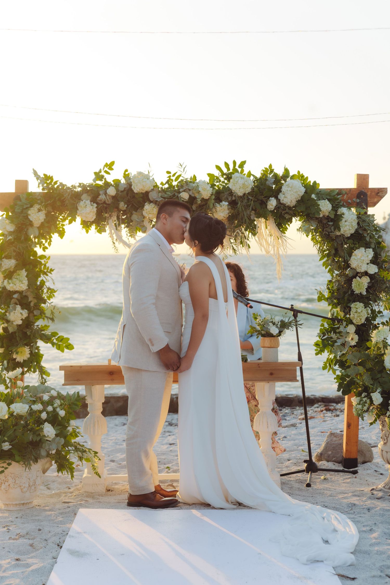 Newlyweds kiss at beach wedding ceremony beneath green and white floral arch at sunset.