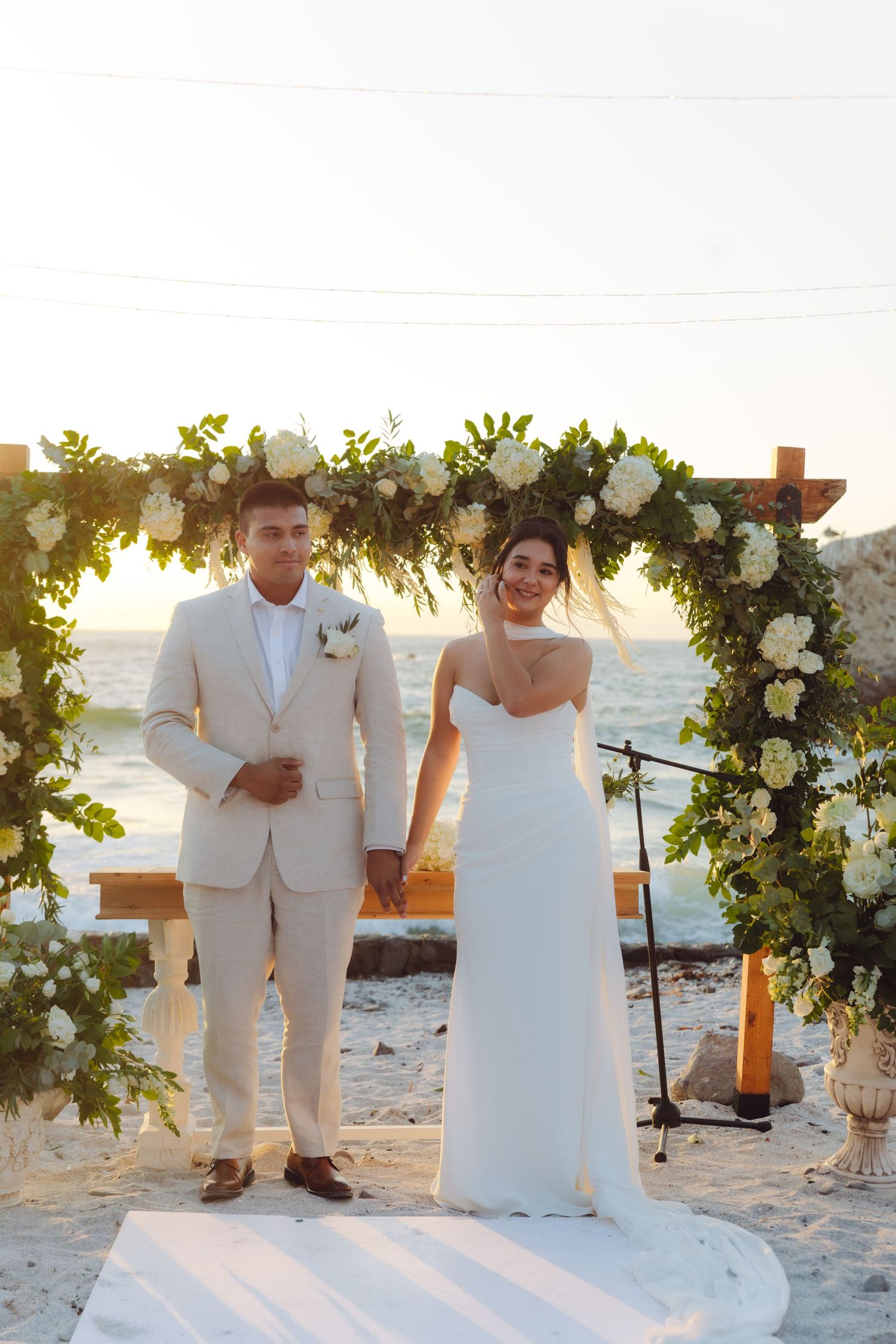Newlyweds pose hand in hand under white floral arch at beach wedding during golden sunset.
