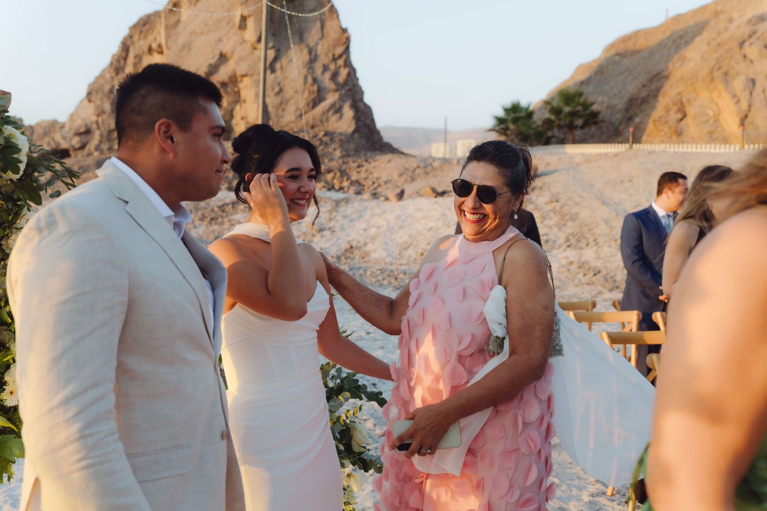 Bride laughing with guest in pink dress at outdoor beach wedding ceremony.