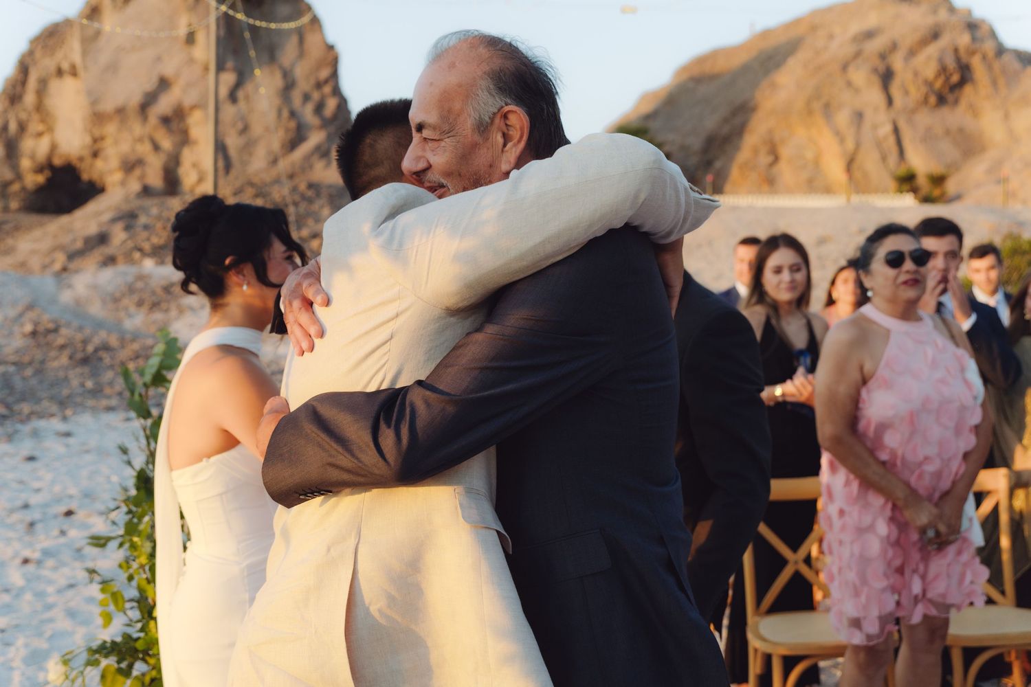 Emotional group hug between bride, groom, and older man at beach wedding.