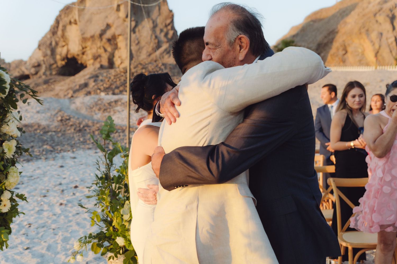 Bride and groom sharing warm embrace with elderly guest at outdoor beach wedding.
