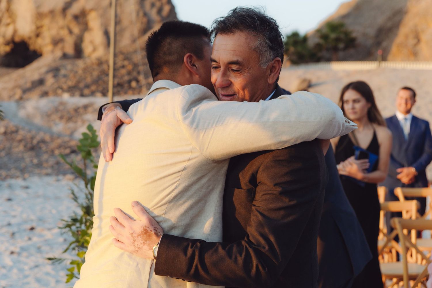 Groom in beige suit embraces older man in warm hug during sunny outdoor beach wedding ceremony.