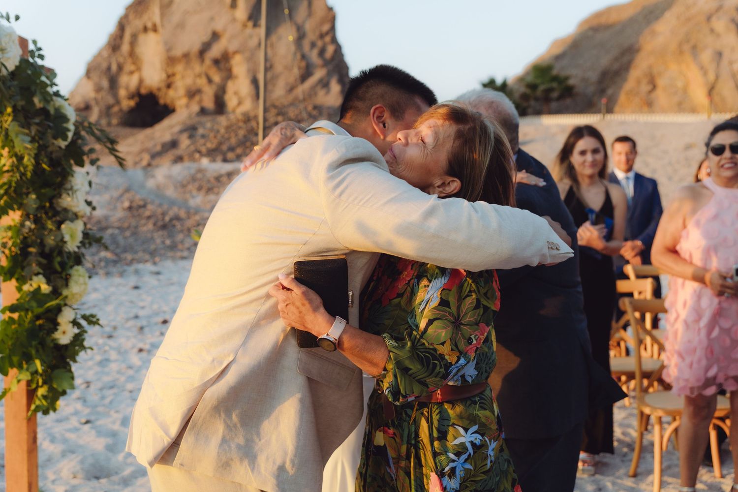 Groom in beige suit shares emotional hug with guest in floral dress during outdoor beach wedding ceremony.