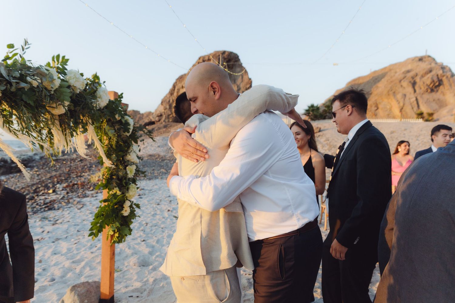 Two men share an emotional embrace under floral arch during outdoor beach wedding ceremony at sunset.
