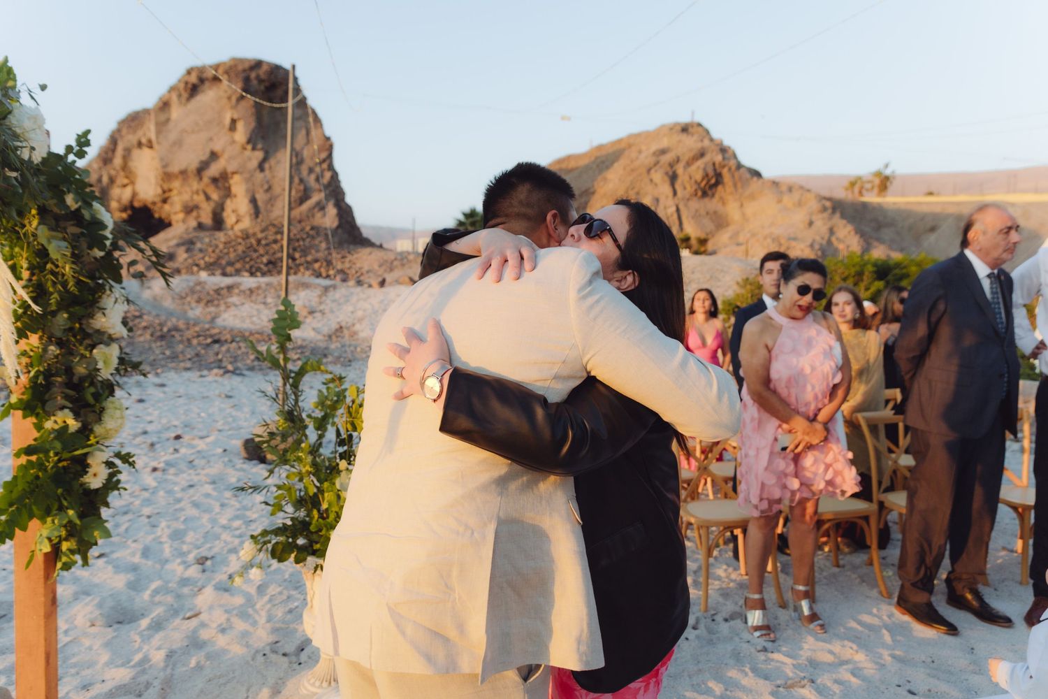 Groom in beige suit shares heartfelt hug with guest during outdoor beach wedding ceremony at golden hour.