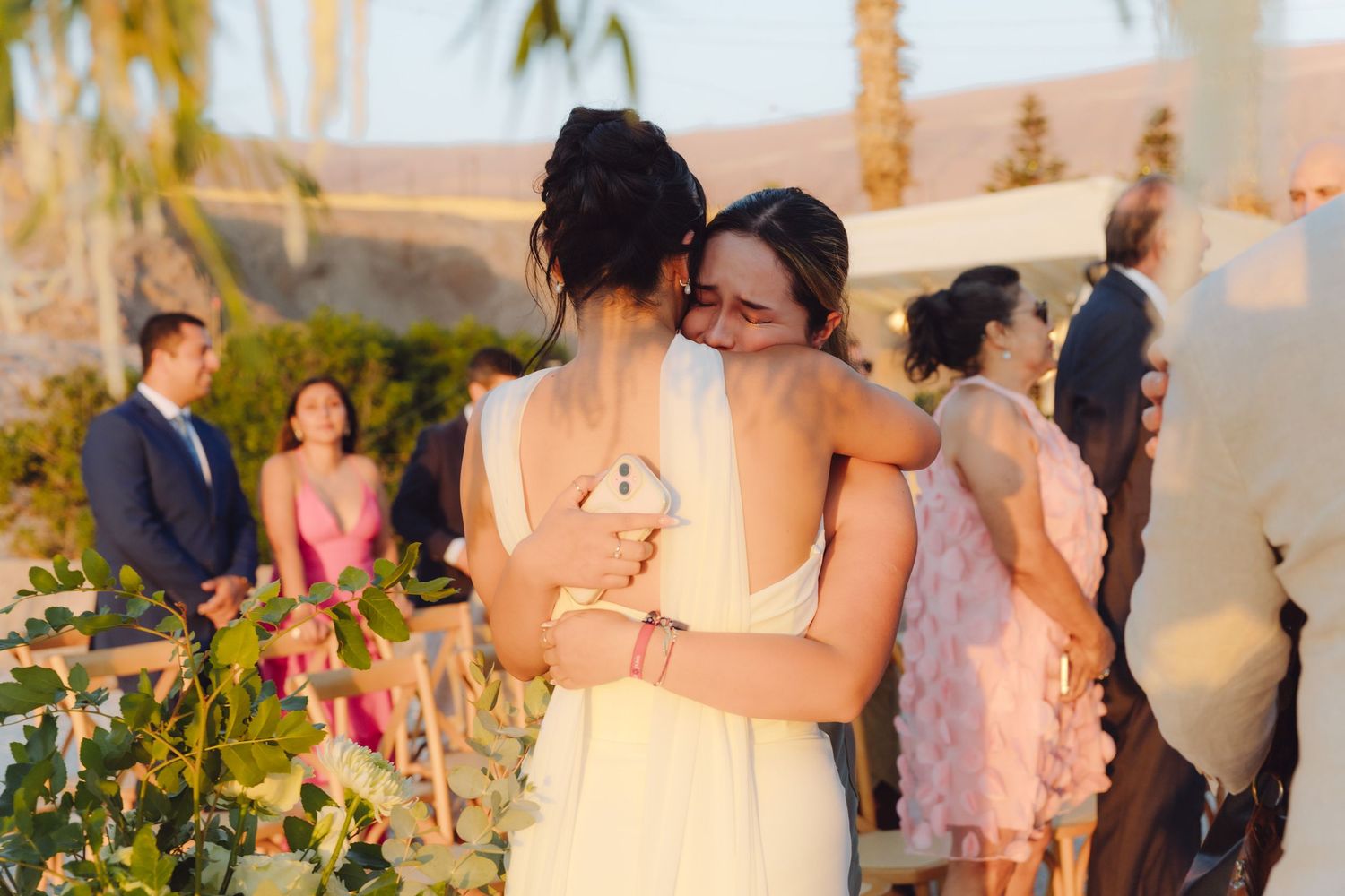 Two women share a warm emotional embrace during outdoor wedding ceremony with guests visible in background.