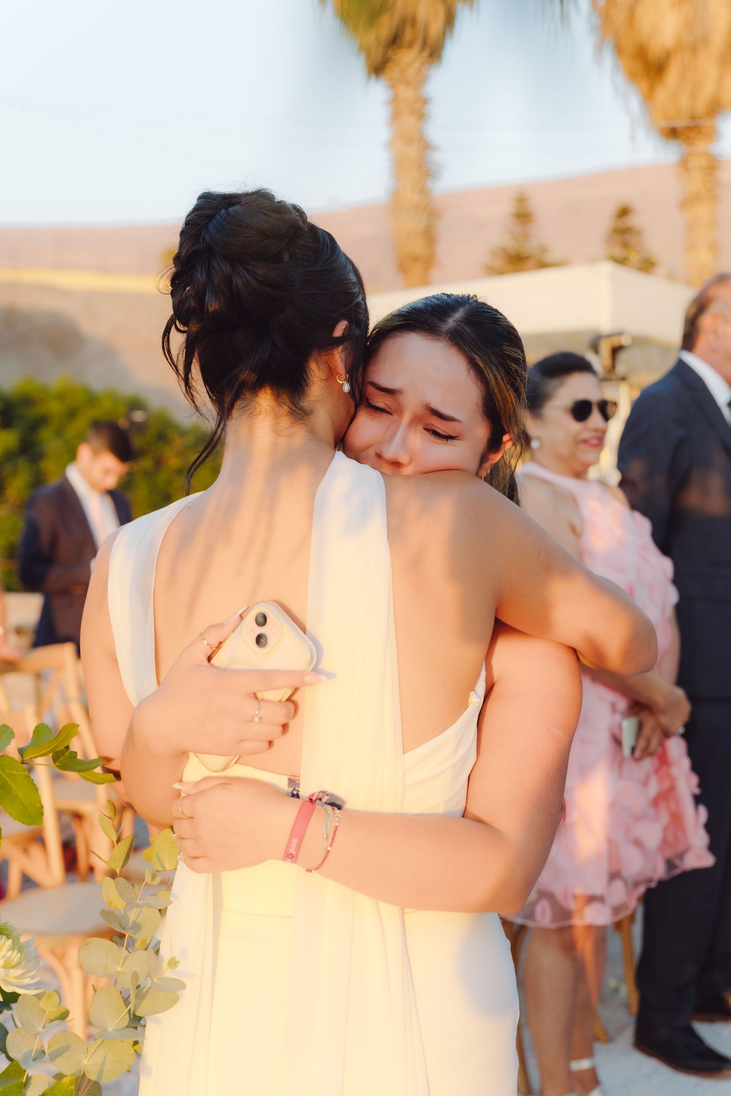 Two women in white dresses share an emotional hug at an outdoor wedding reception with palm trees.
