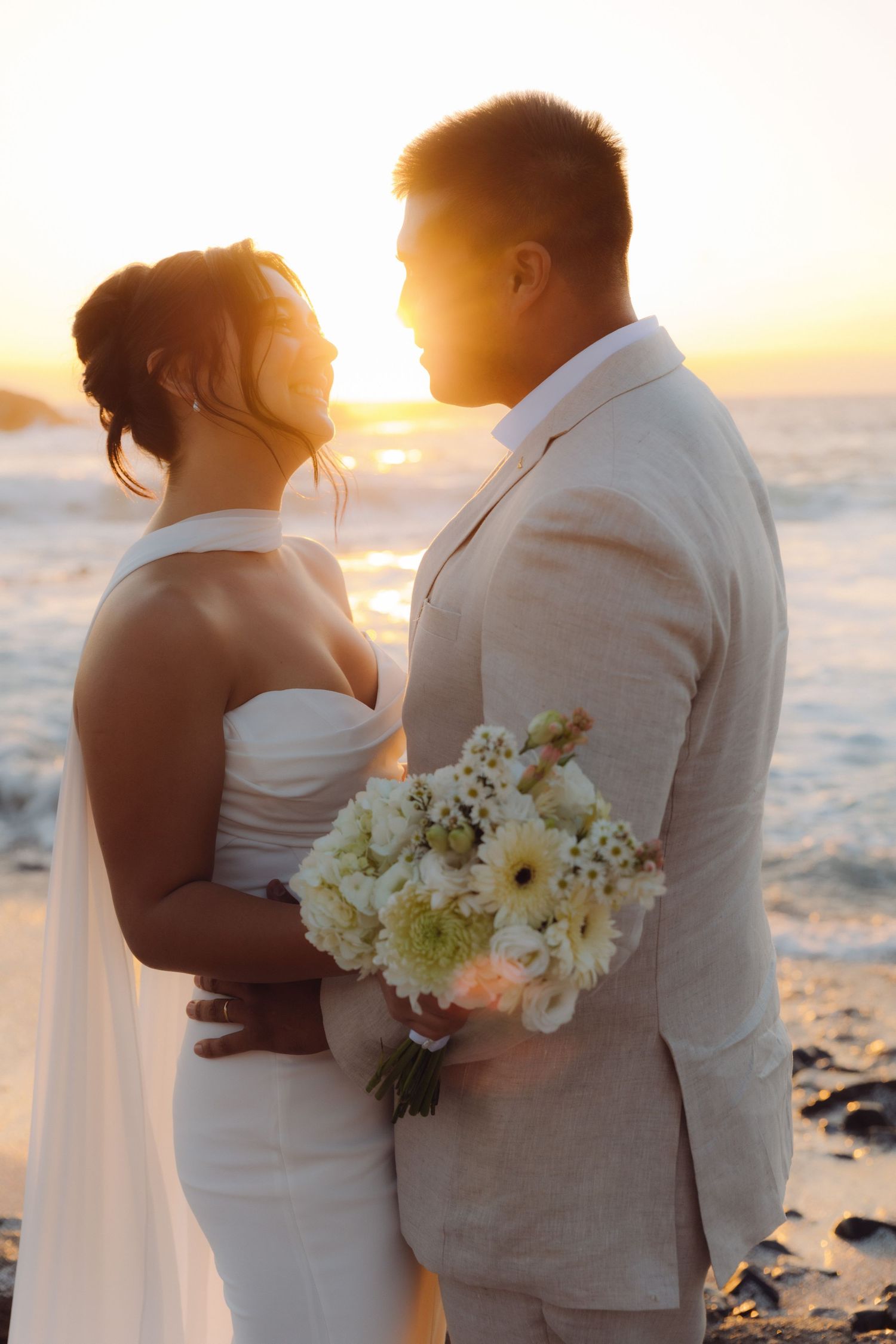 Bride and groom embrace at sunset on a rocky beach, holding white flower bouquet.
