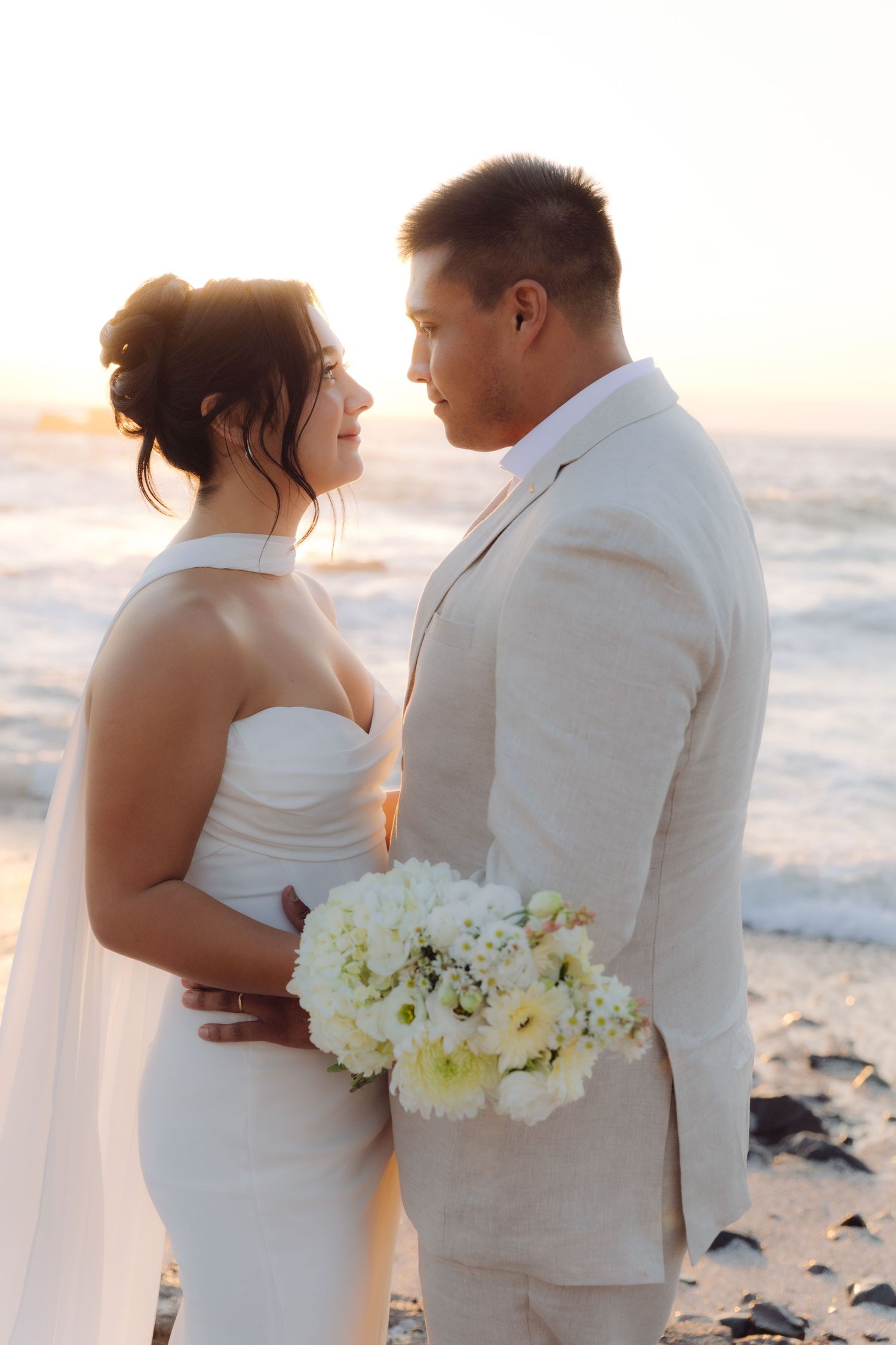 Wedding couple stands face to face at beach sunset, bride holding white floral bouquet.