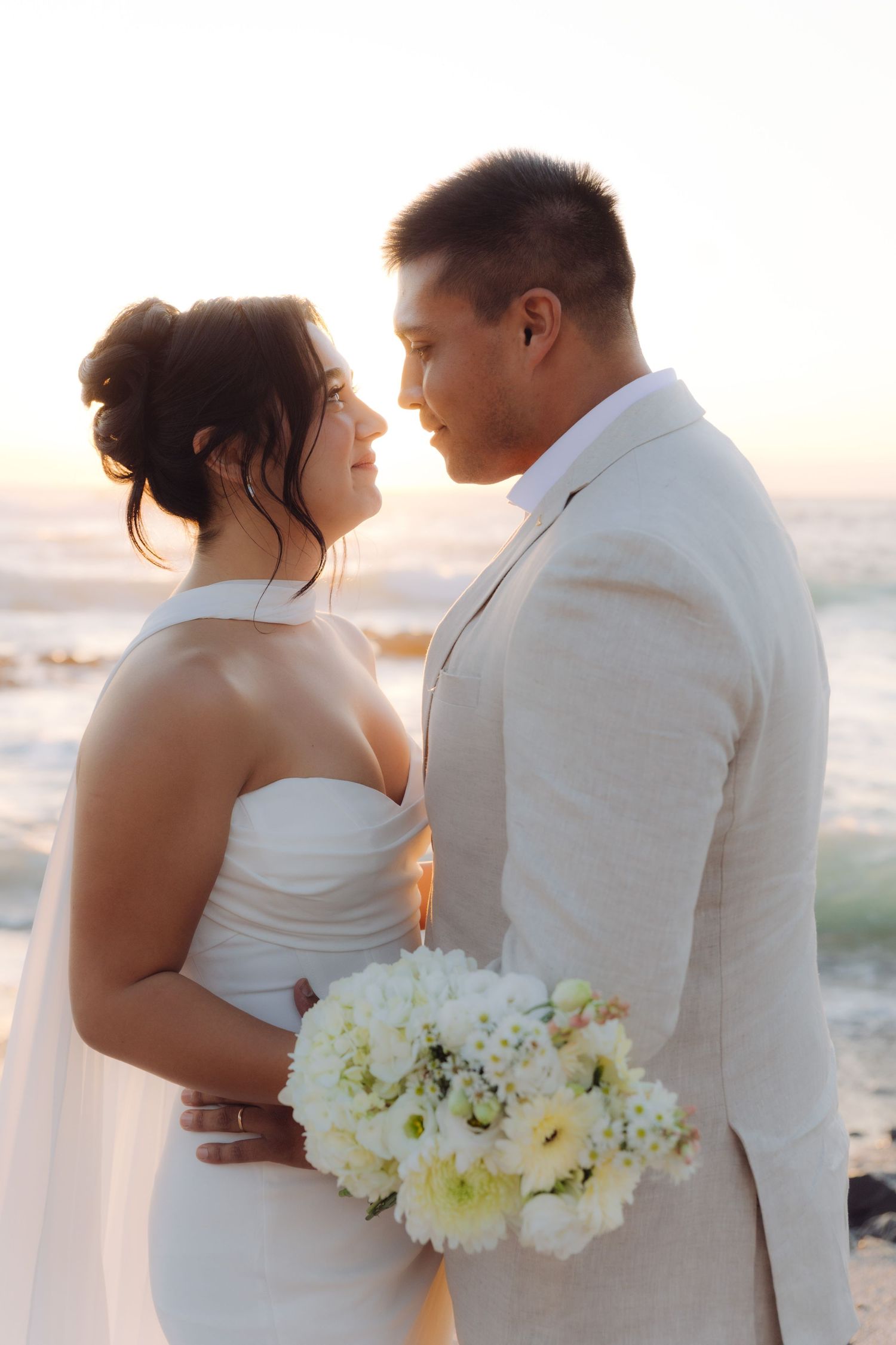 Newlyweds share intimate moment at golden hour beach wedding, groom holding white bouquet.