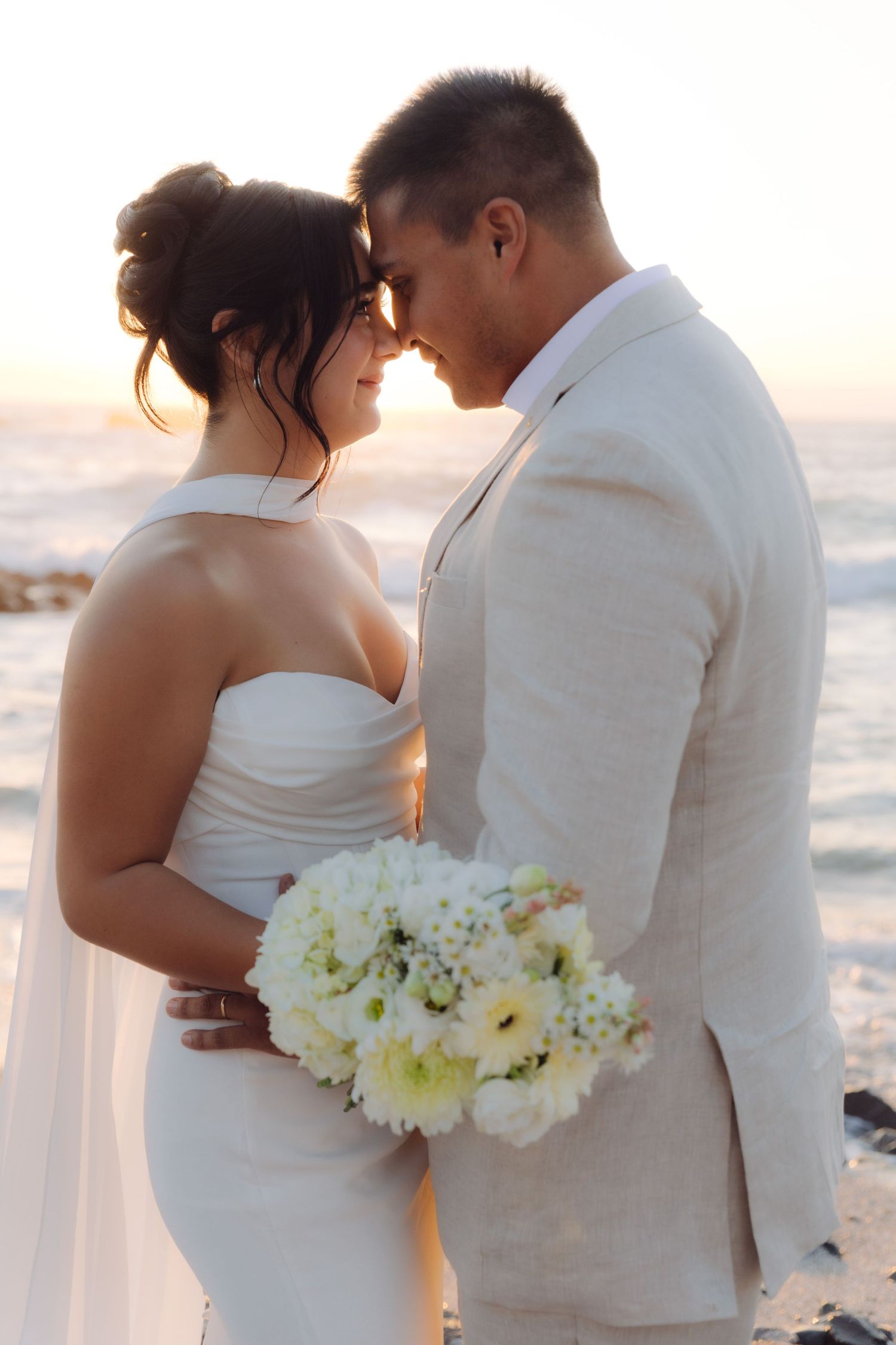 Bride and groom touch foreheads romantically at sunset beach wedding with white bouquet.