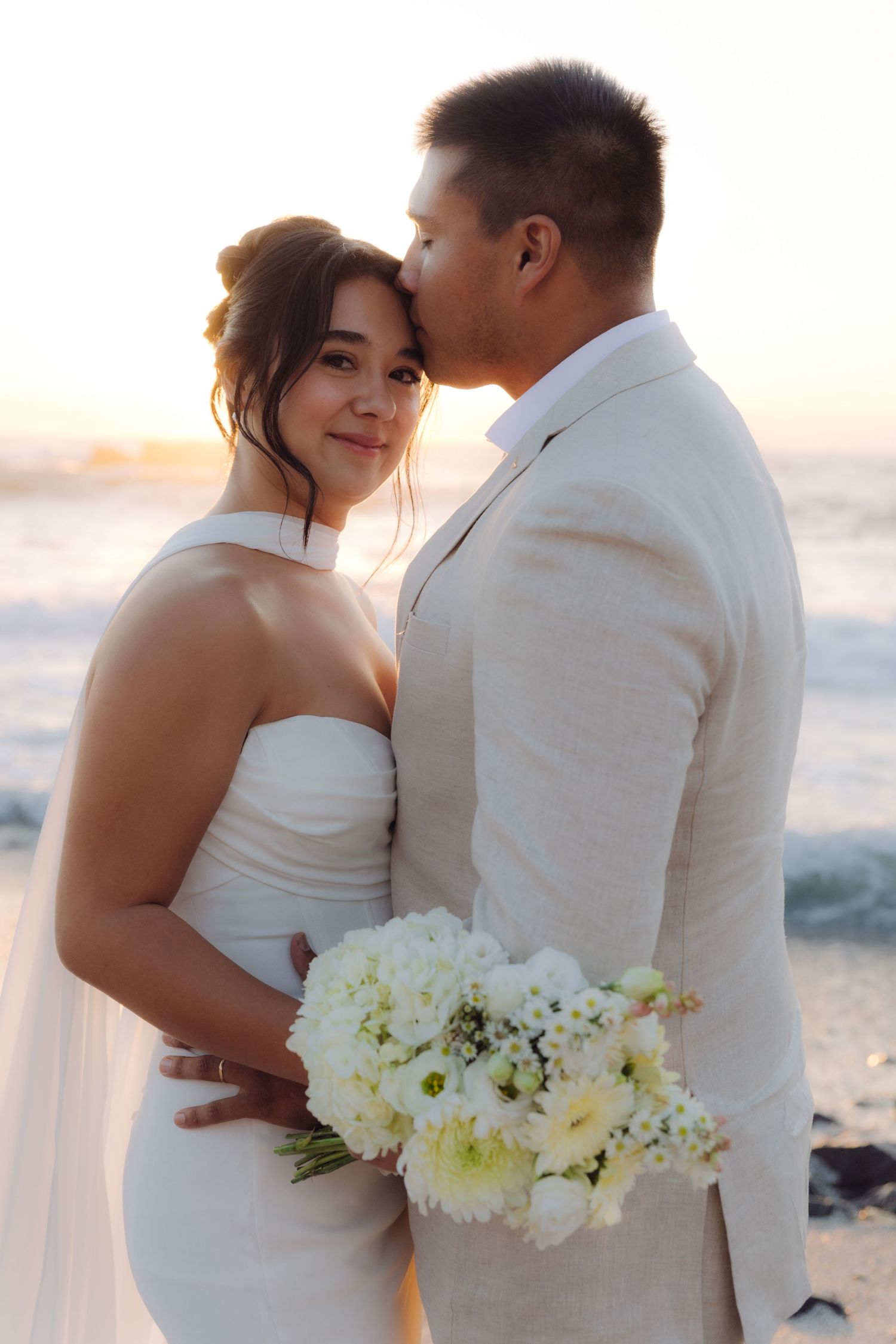 Groom kisses smiling bride on forehead at golden sunset beach wedding ceremony.