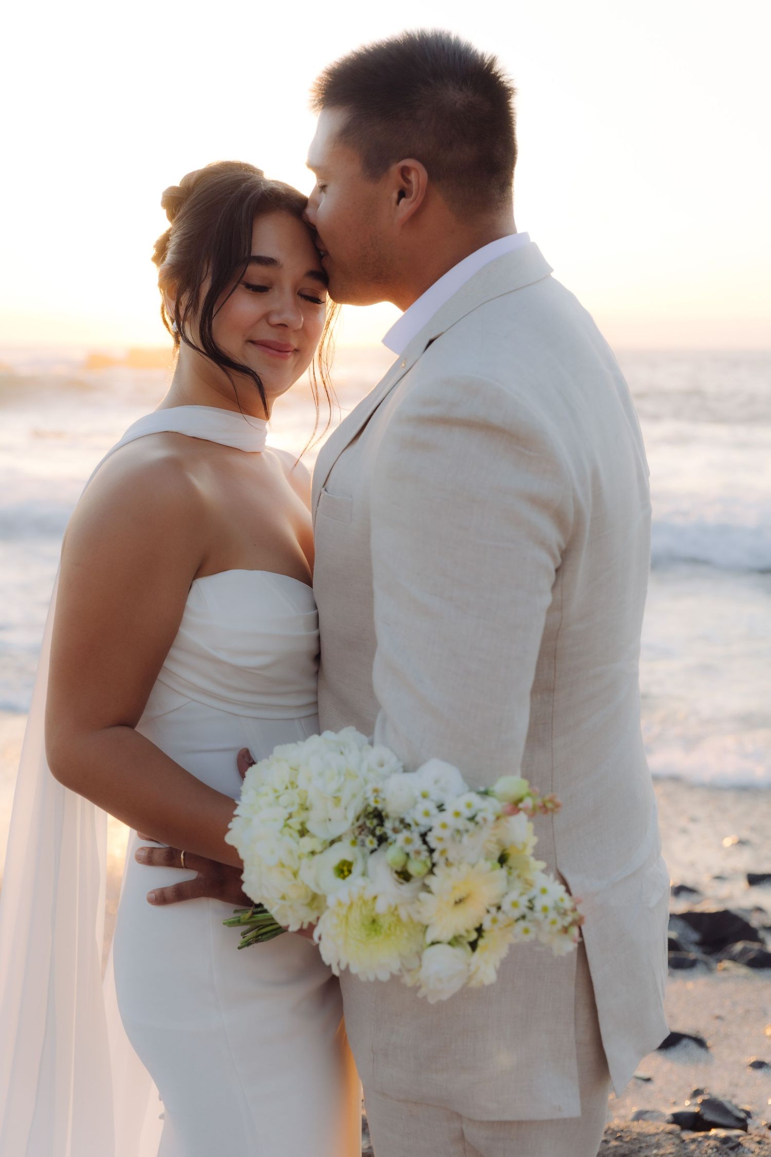 Groom tenderly kisses bride on cheek at romantic beach wedding during sunset.