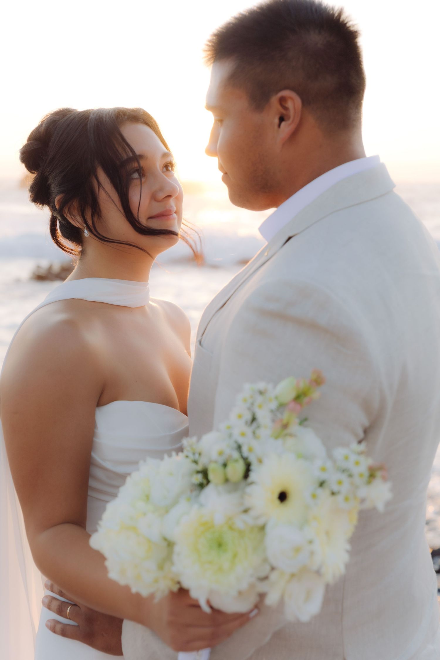 Smiling bride gazes at groom during golden hour beach wedding holding white bouquet.