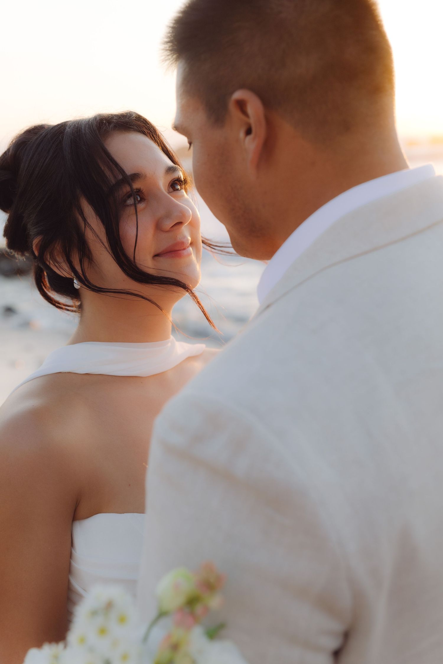 Bride looks lovingly up at groom during intimate beach wedding portrait at sunset.