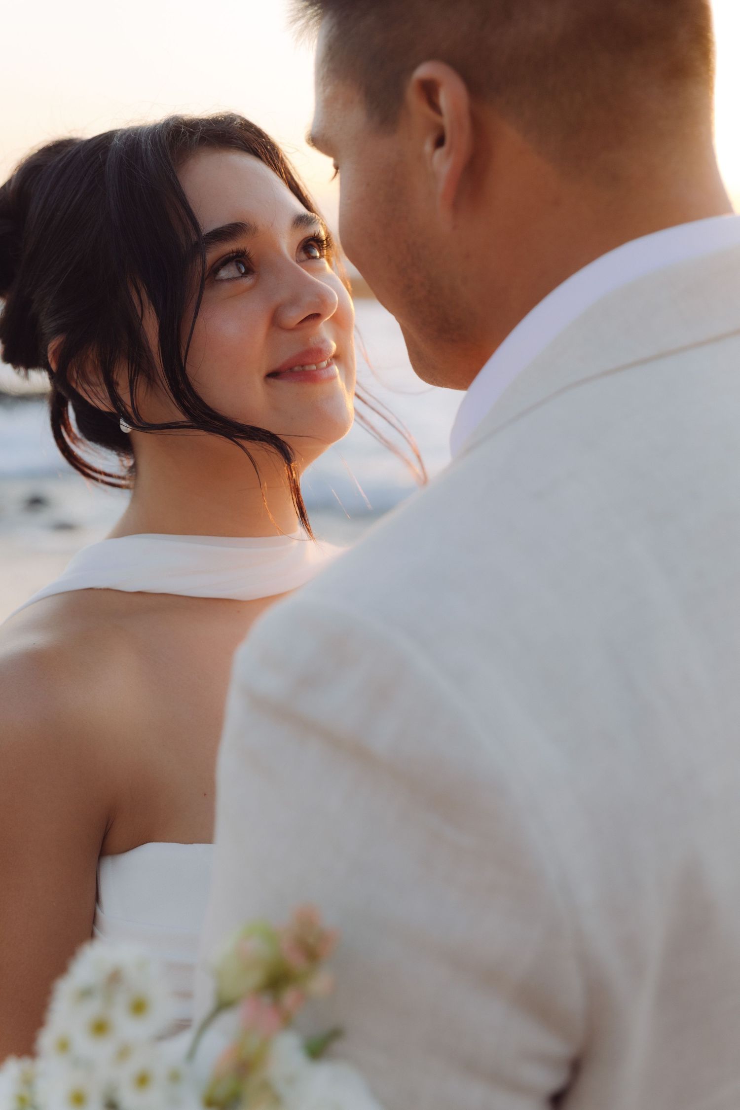 Bride smiles up at groom during romantic beach wedding portrait at golden sunset.