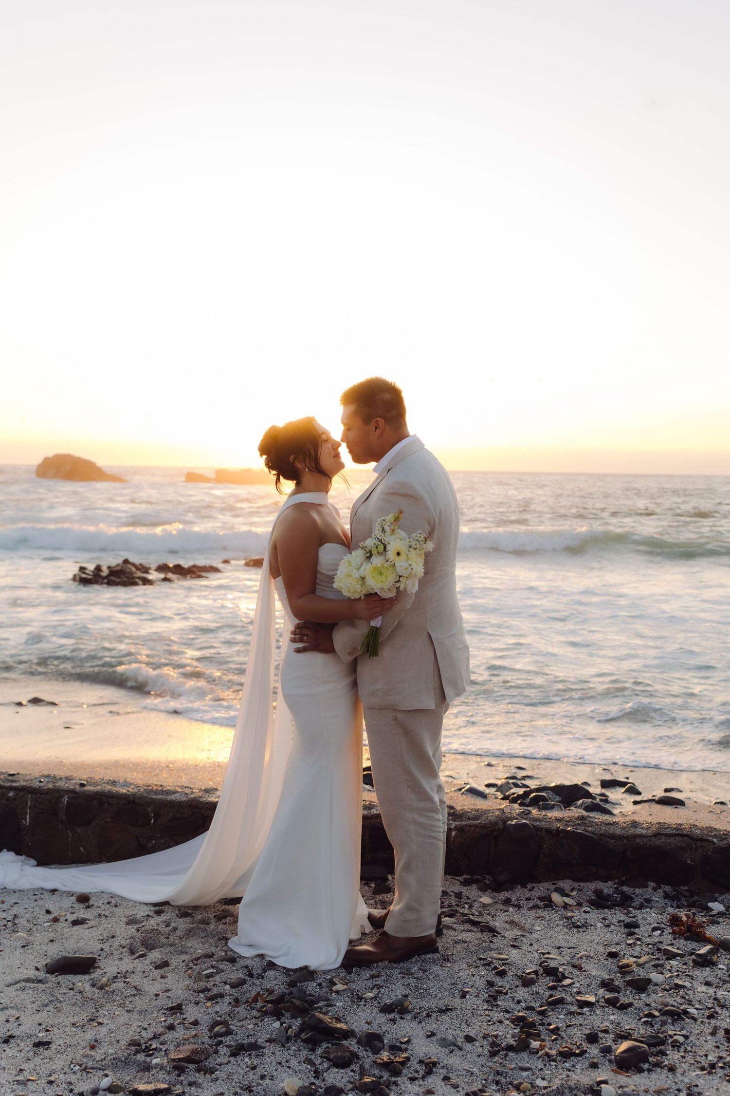 Bride and groom share a romantic kiss on a rocky beach at sunset, bride holding white bouquet.