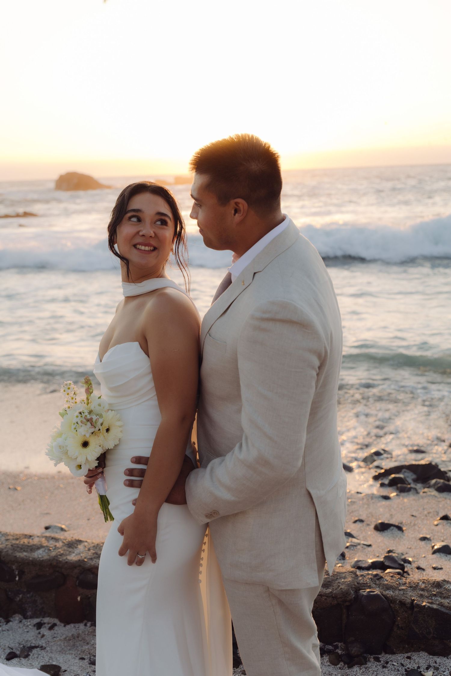 Smiling bride in white halter gown holds bouquet as groom embraces her at ocean sunset.