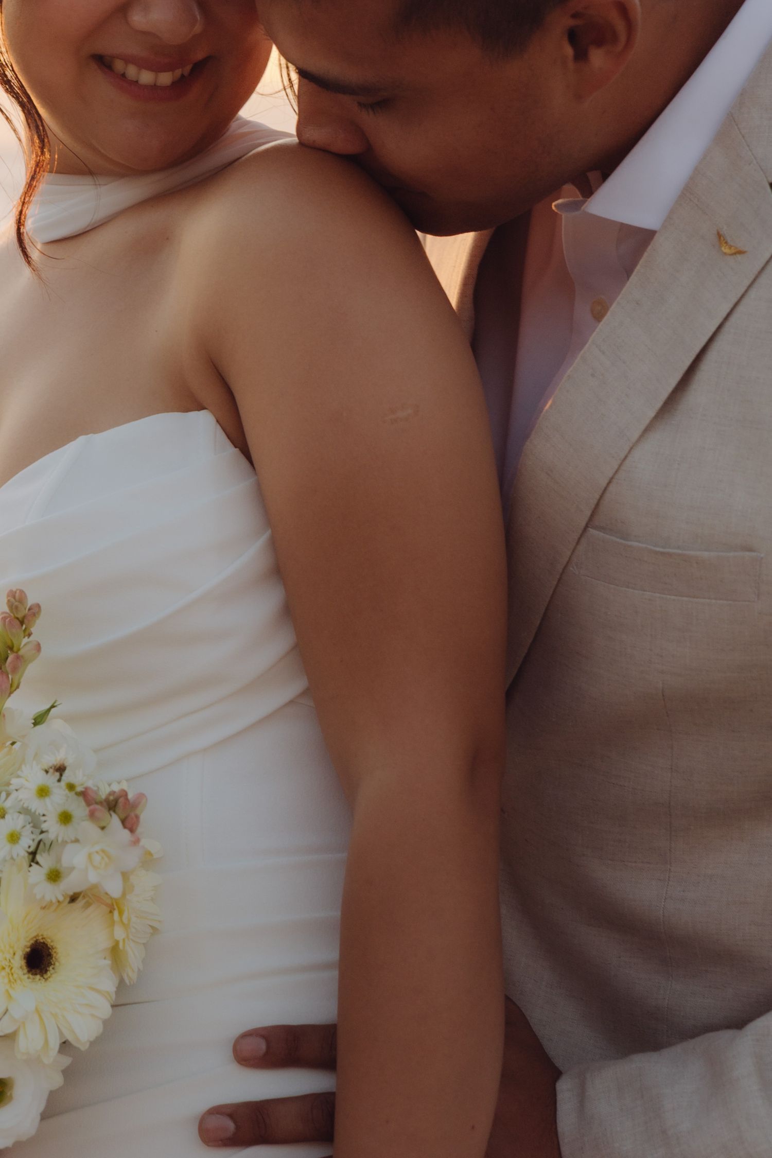 Close-up of groom in beige suit kissing bride's shoulder while she holds white gerbera bouquet.
