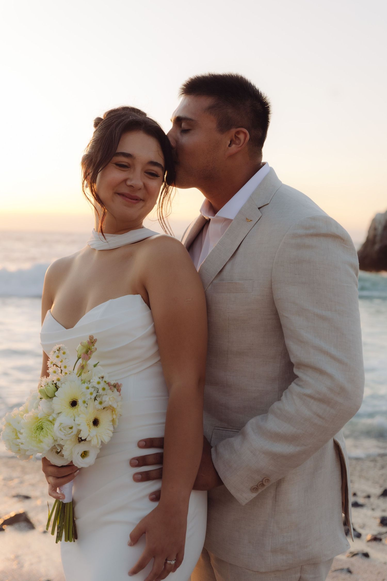 Groom in tan suit kisses bride's temple as she smiles holding white floral bouquet at beach sunset.
