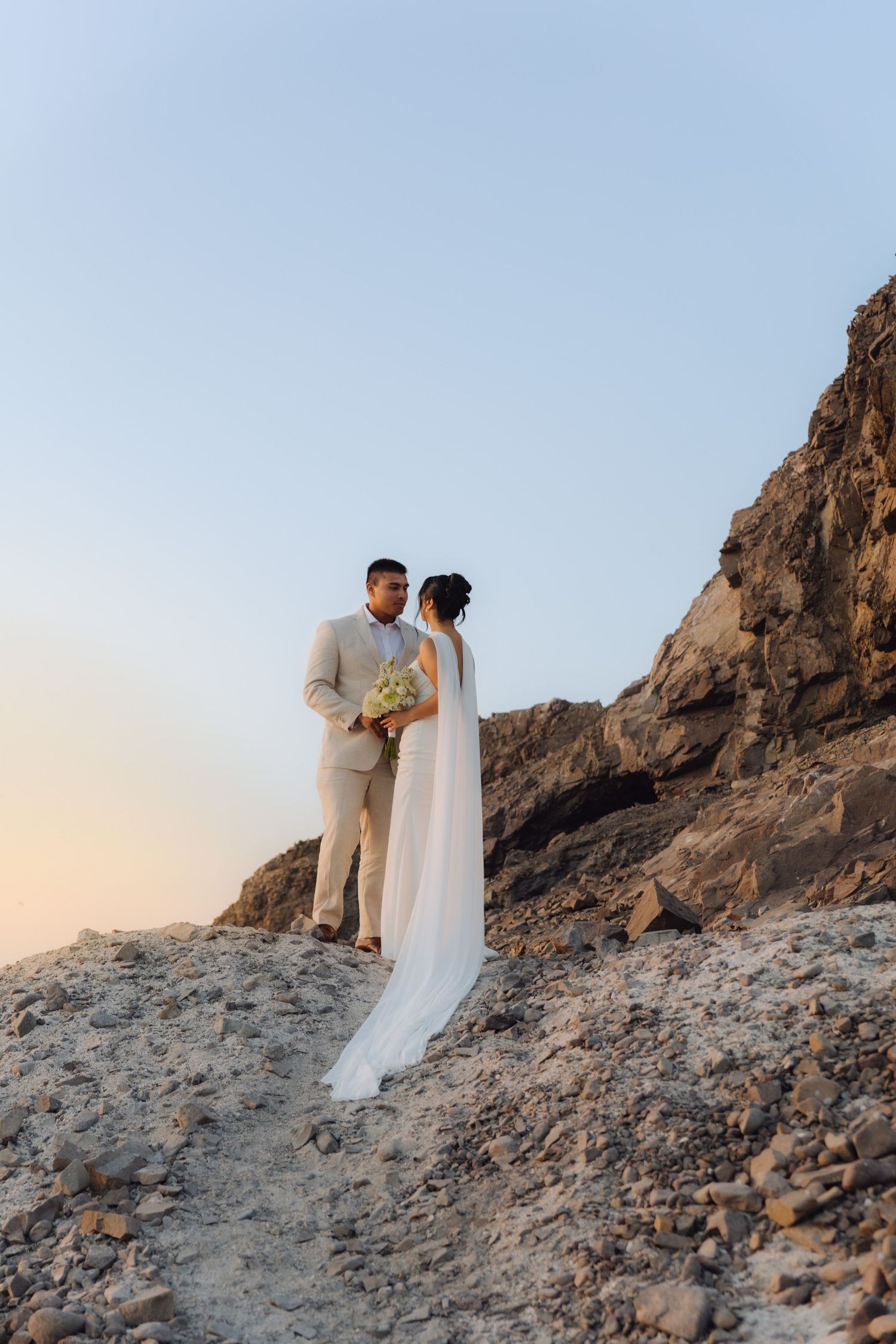 Bride and groom stand together on rocky coastal cliffside at dusk, bride wearing flowing white veil.