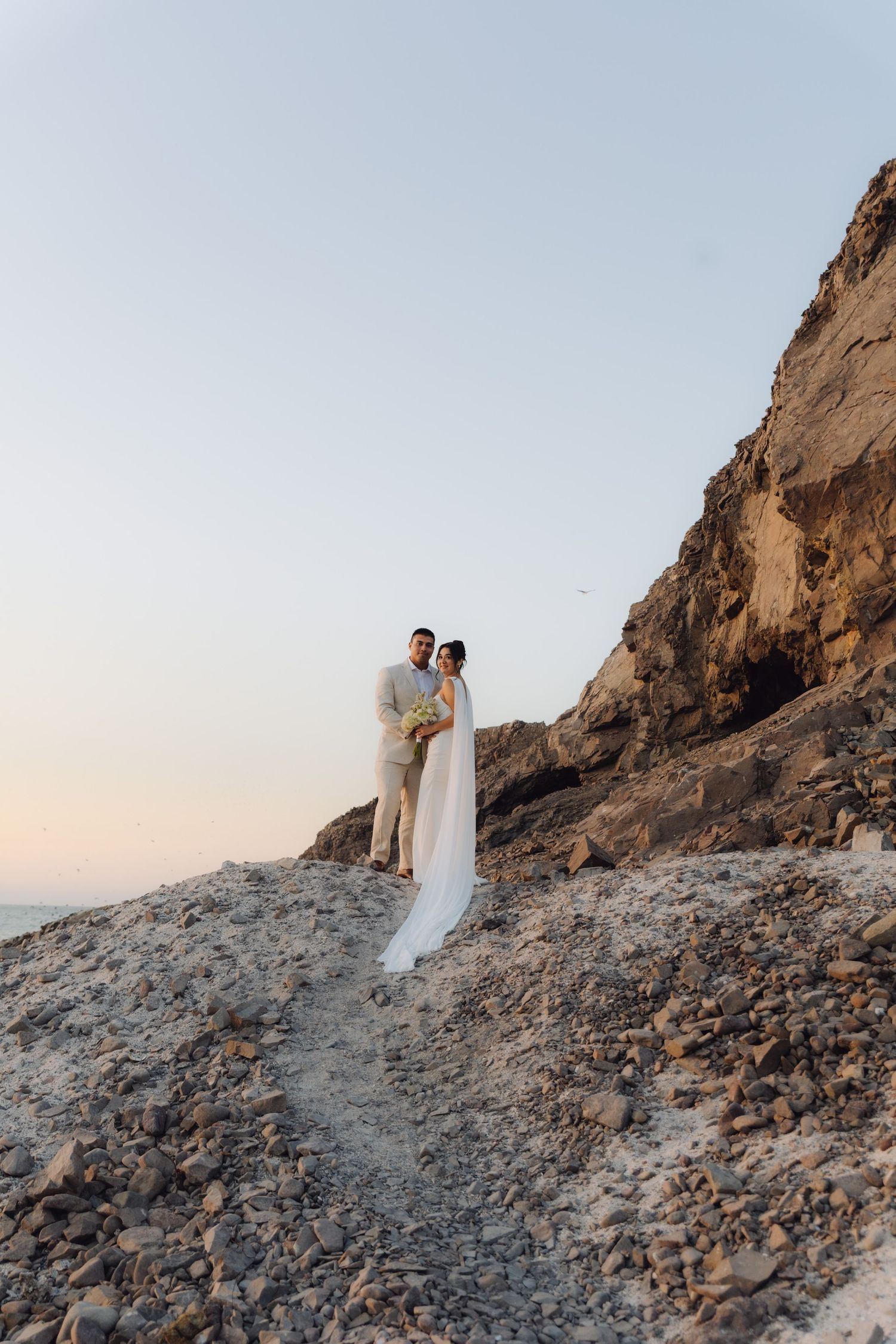 Wedding couple embraces on rocky beach cliff at sunset, bride in white gown with long flowing veil.
