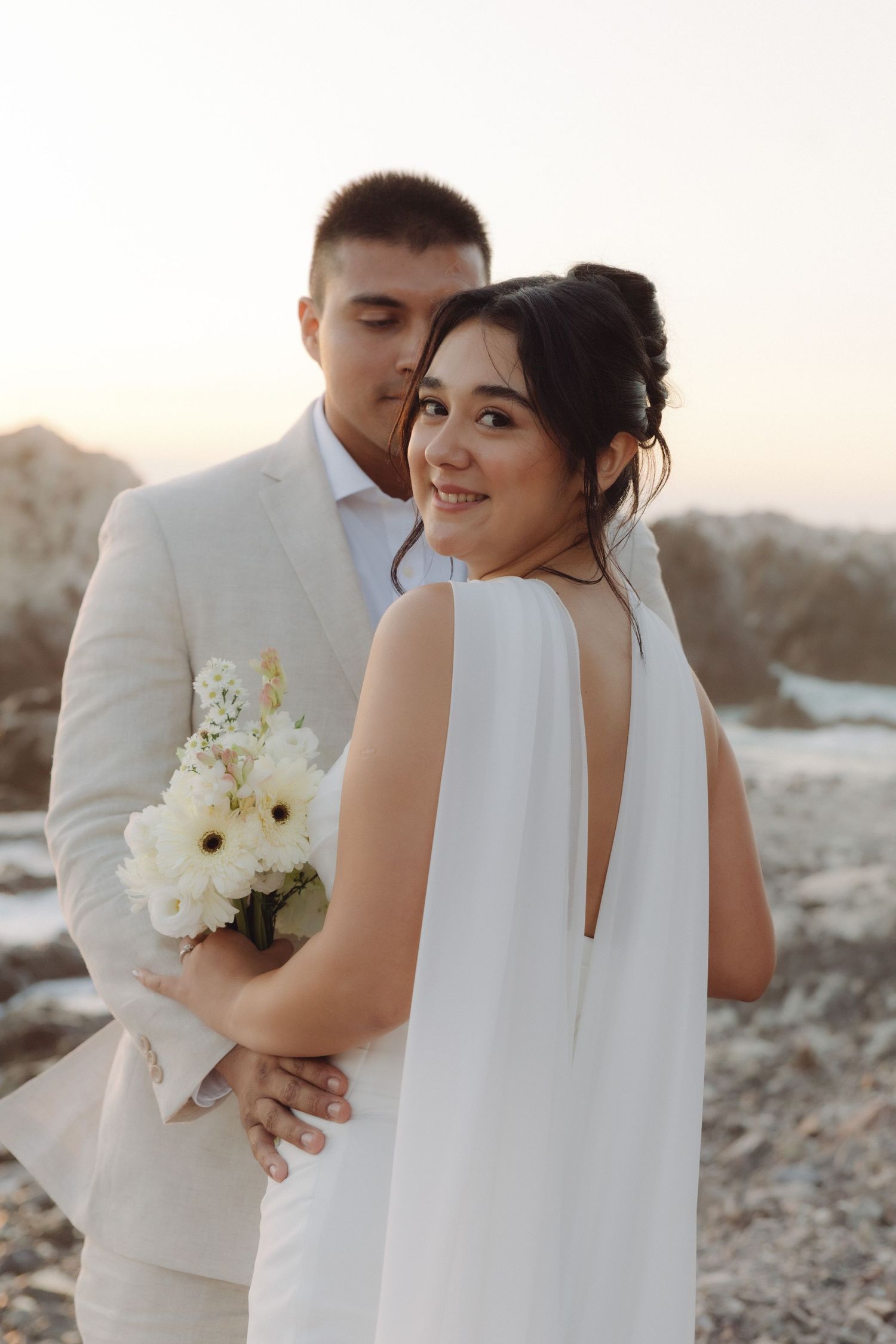 Bride in white cape-back gown smiles over shoulder as groom embraces her at rocky sunset beach.