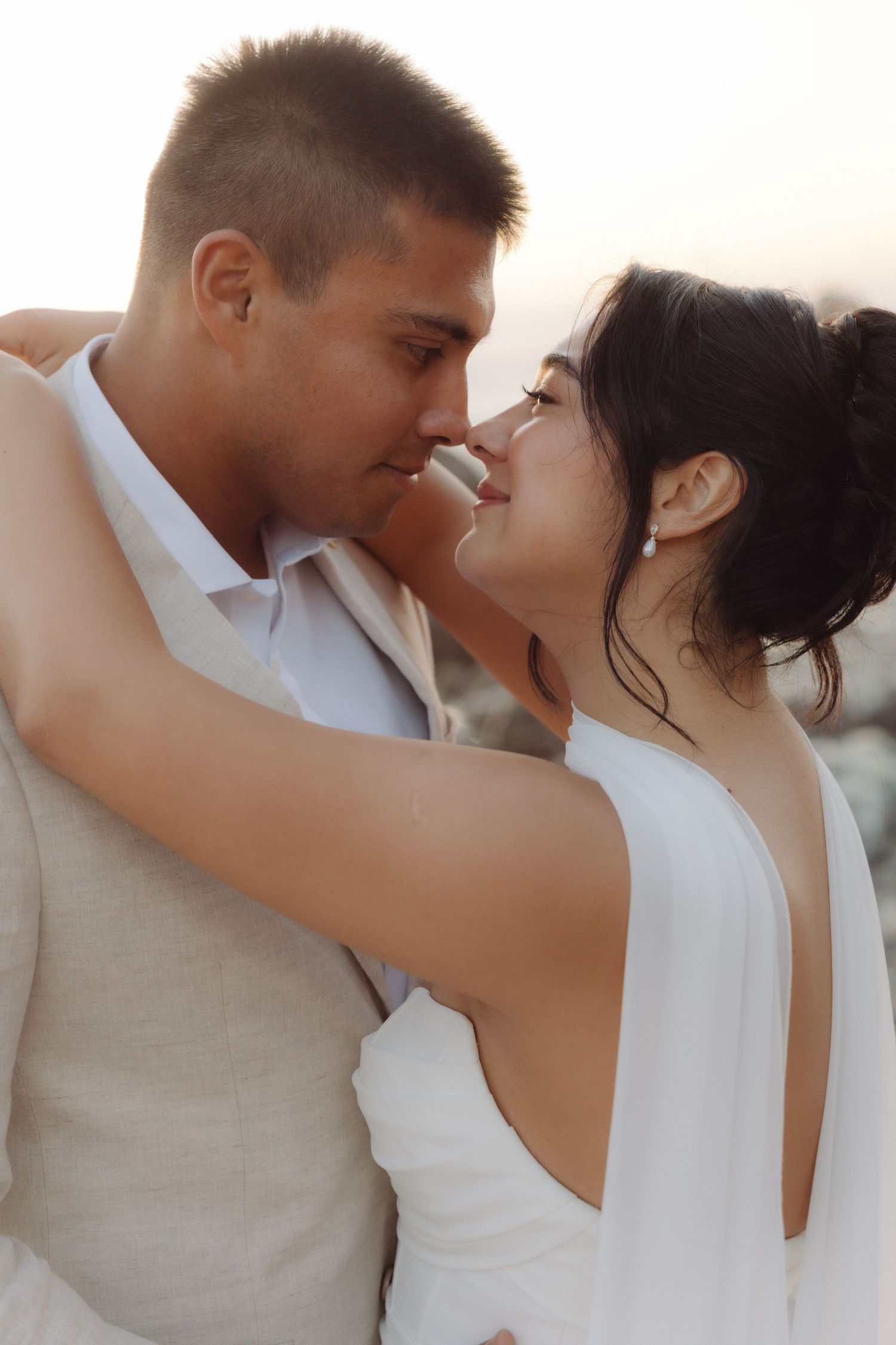 Bride and groom lean in for a kiss, embracing closely against a soft sunset sky backdrop.