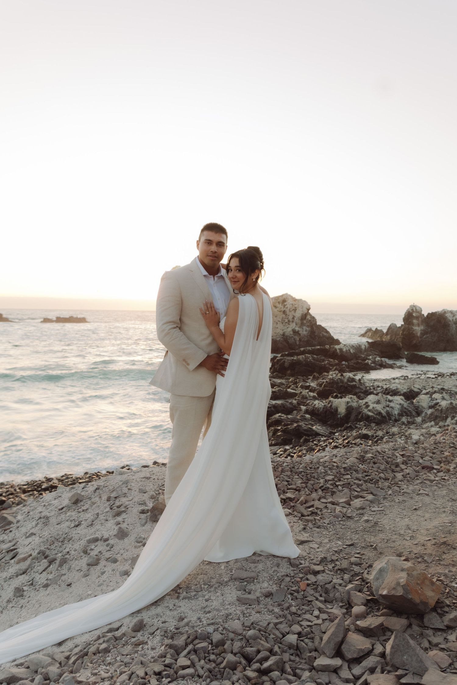 Groom in beige suit holds bride in white open-back gown near rocky ocean shore at sunset.