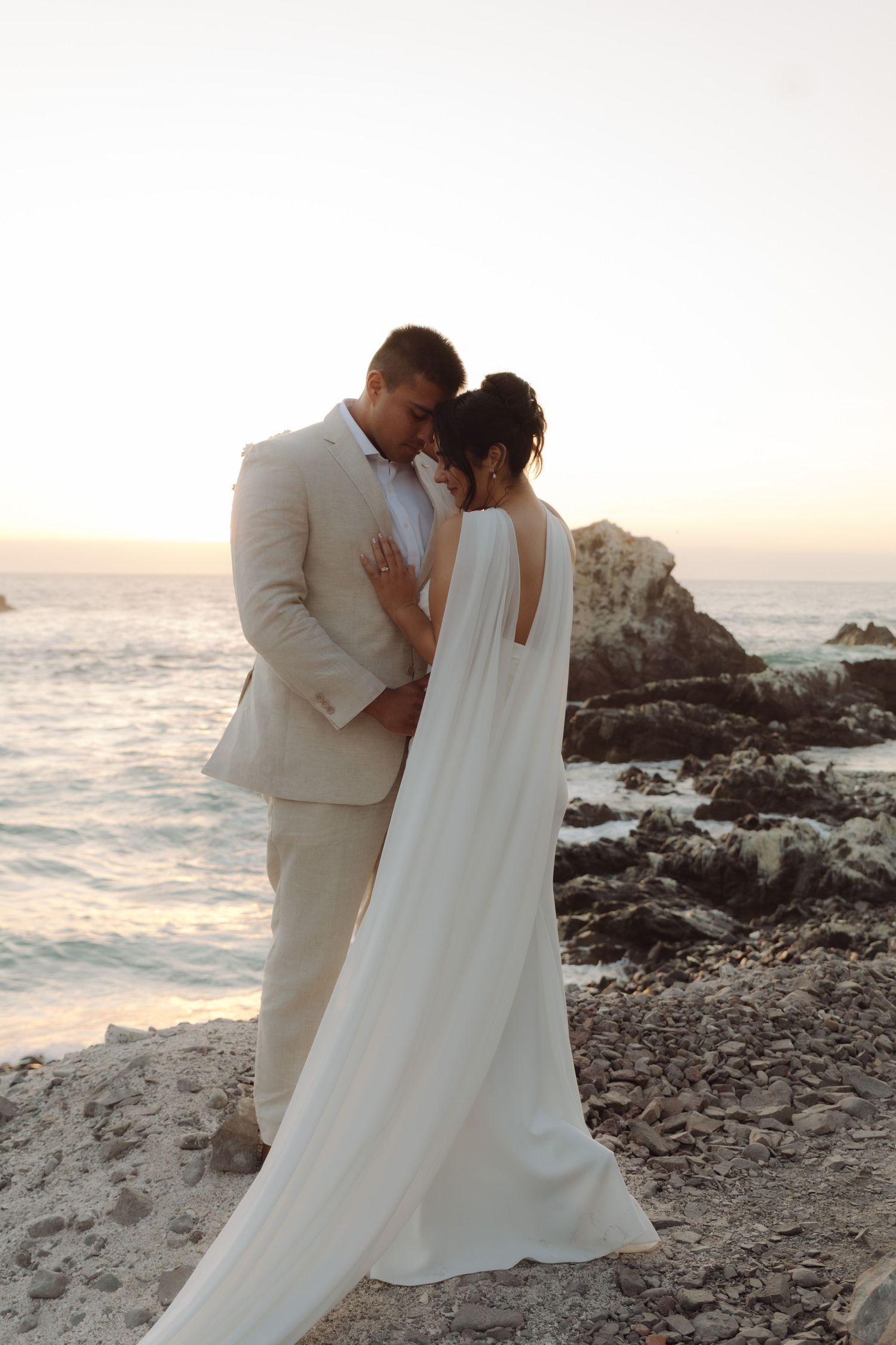 Bride and groom share an intimate forehead-to-forehead moment on rocky beach at golden sunset.