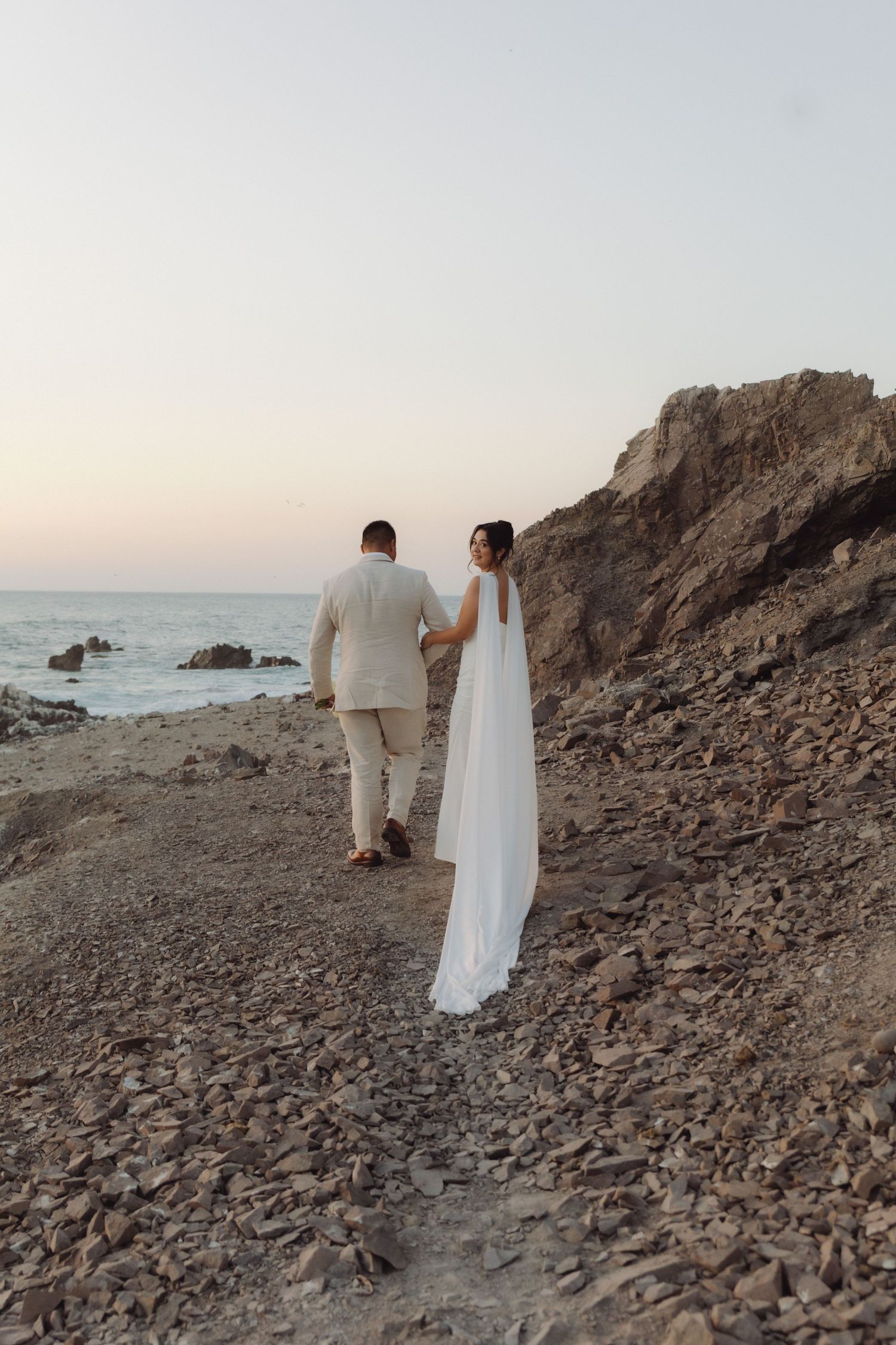Newlywed couple walks hand in hand along a scenic rocky coastline at dusk, bride in elegant white dress with cape veil.