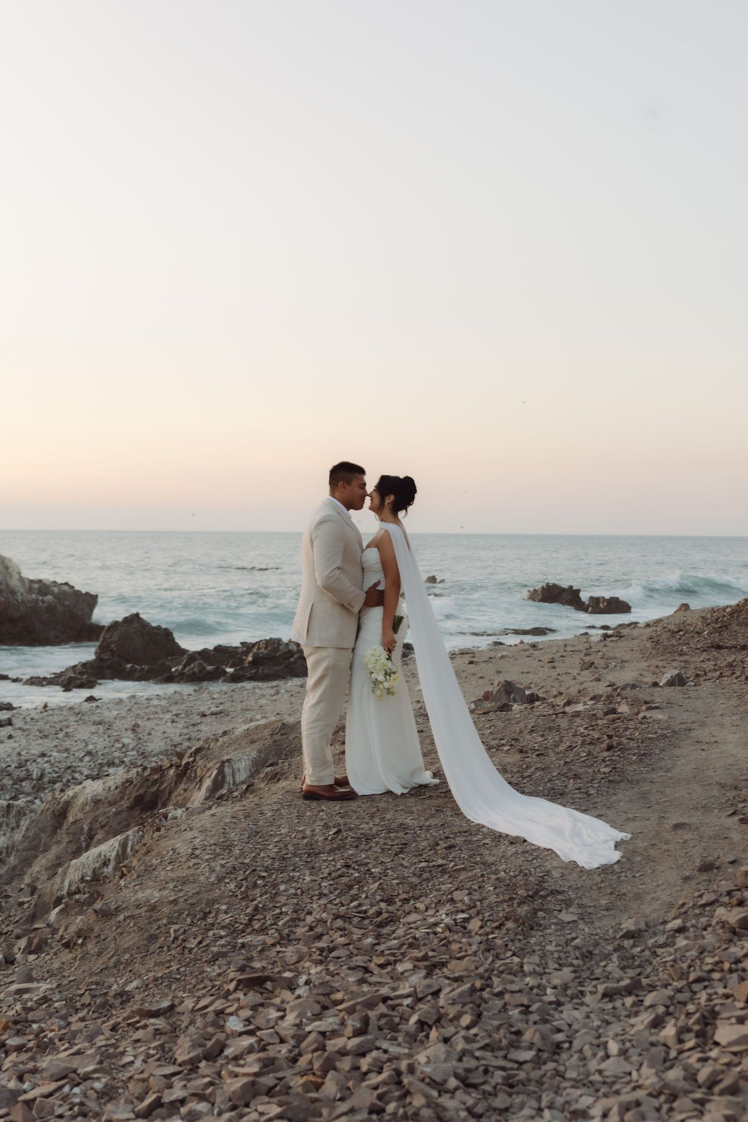 Bride and groom share a romantic moment on a rocky beach at sunset, bride wearing a flowing white gown with long veil.