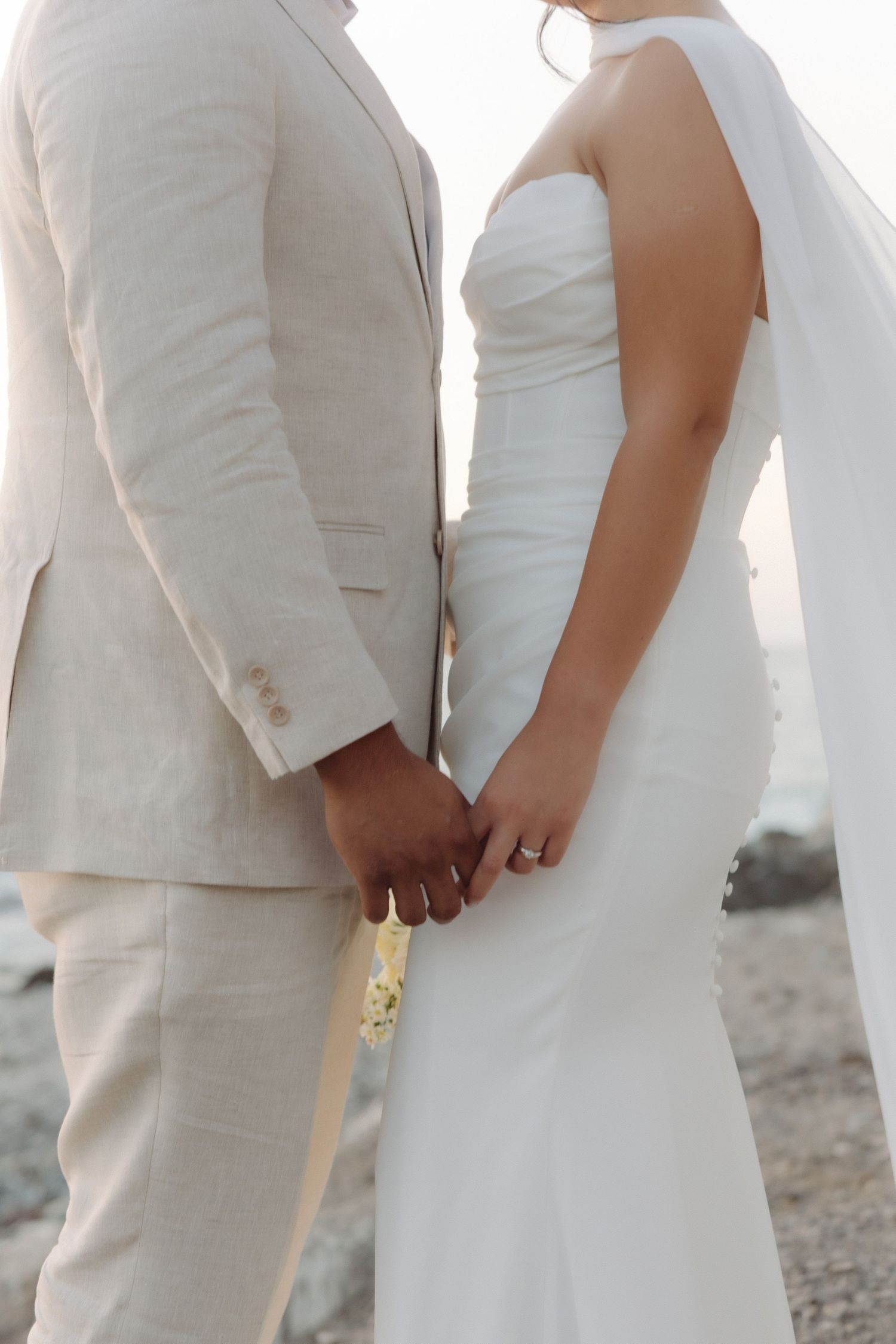 Close-up of bride and groom holding hands at their beach wedding, bride wearing a white strapless gown and diamond ring.