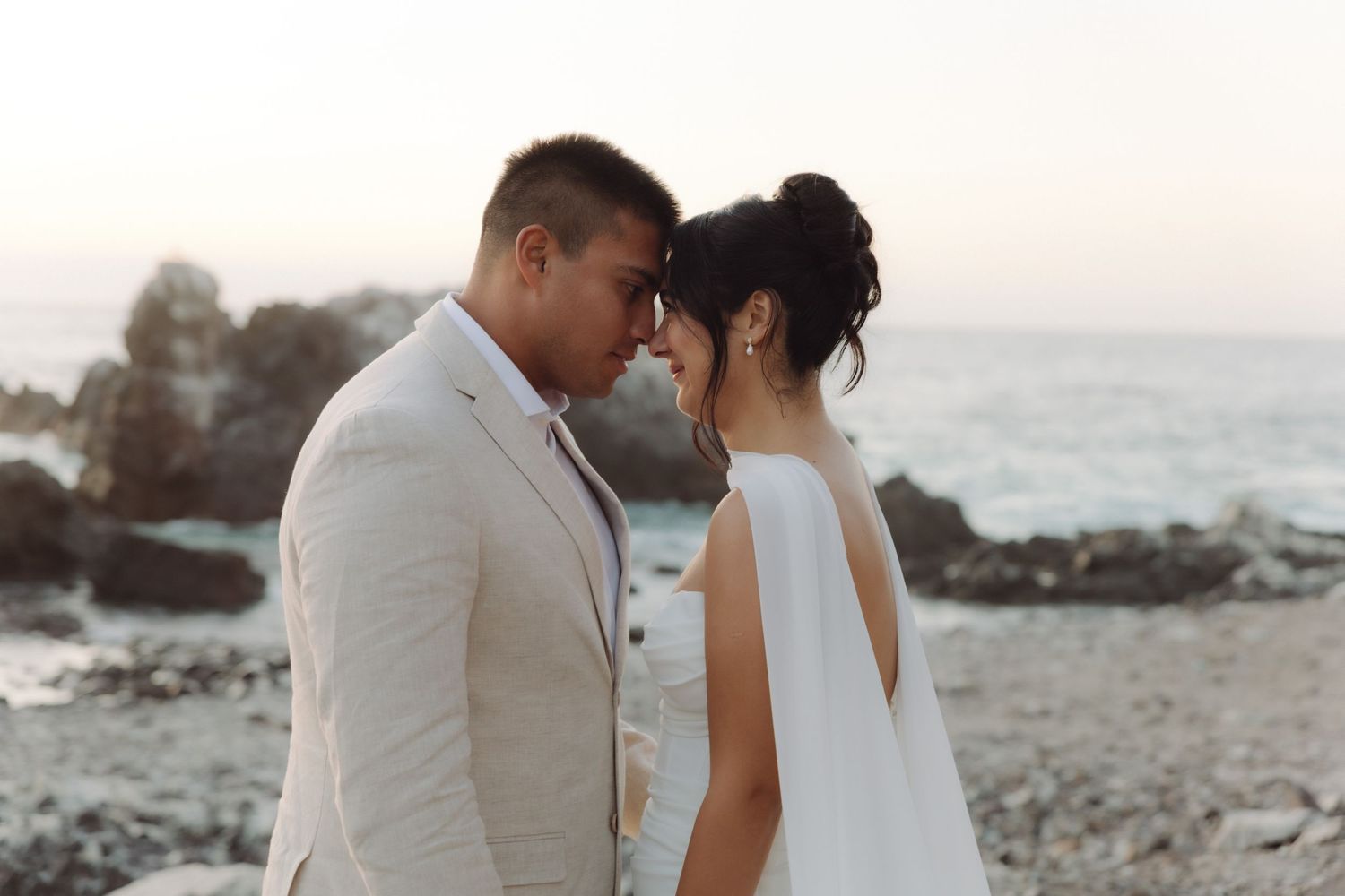 Bride and groom touch foreheads in an intimate moment on a rocky beach at sunset, ocean waves in the background.
