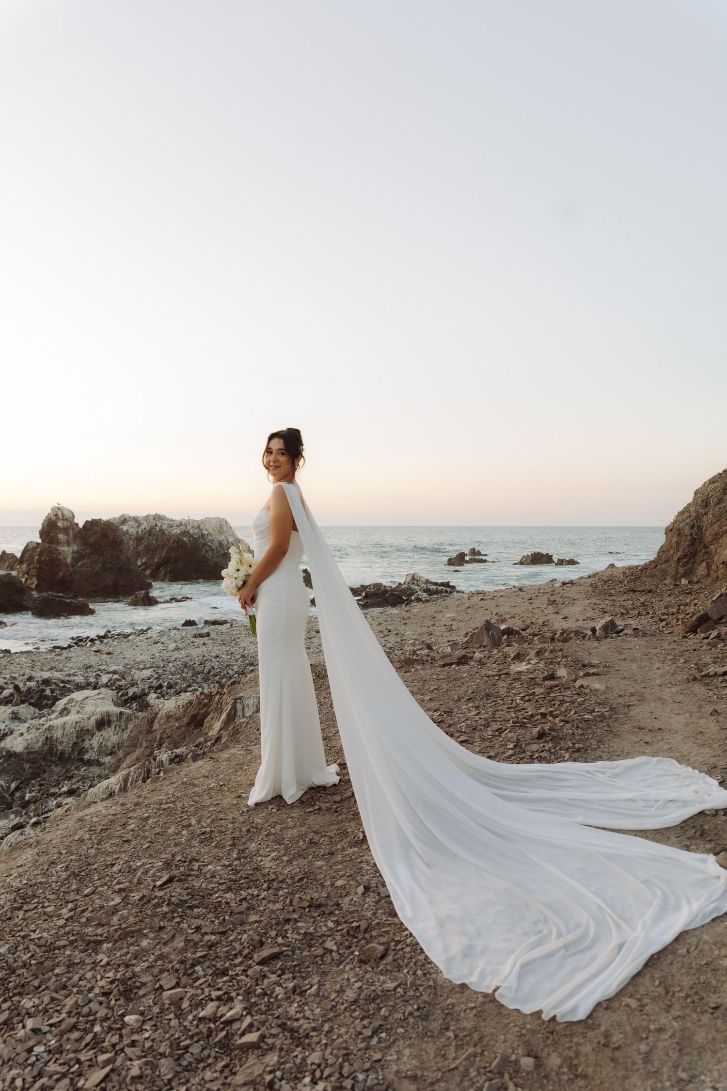 Elegant bride in a white halter gown with dramatic cathedral veil poses on a rocky beach at sunset by the ocean.