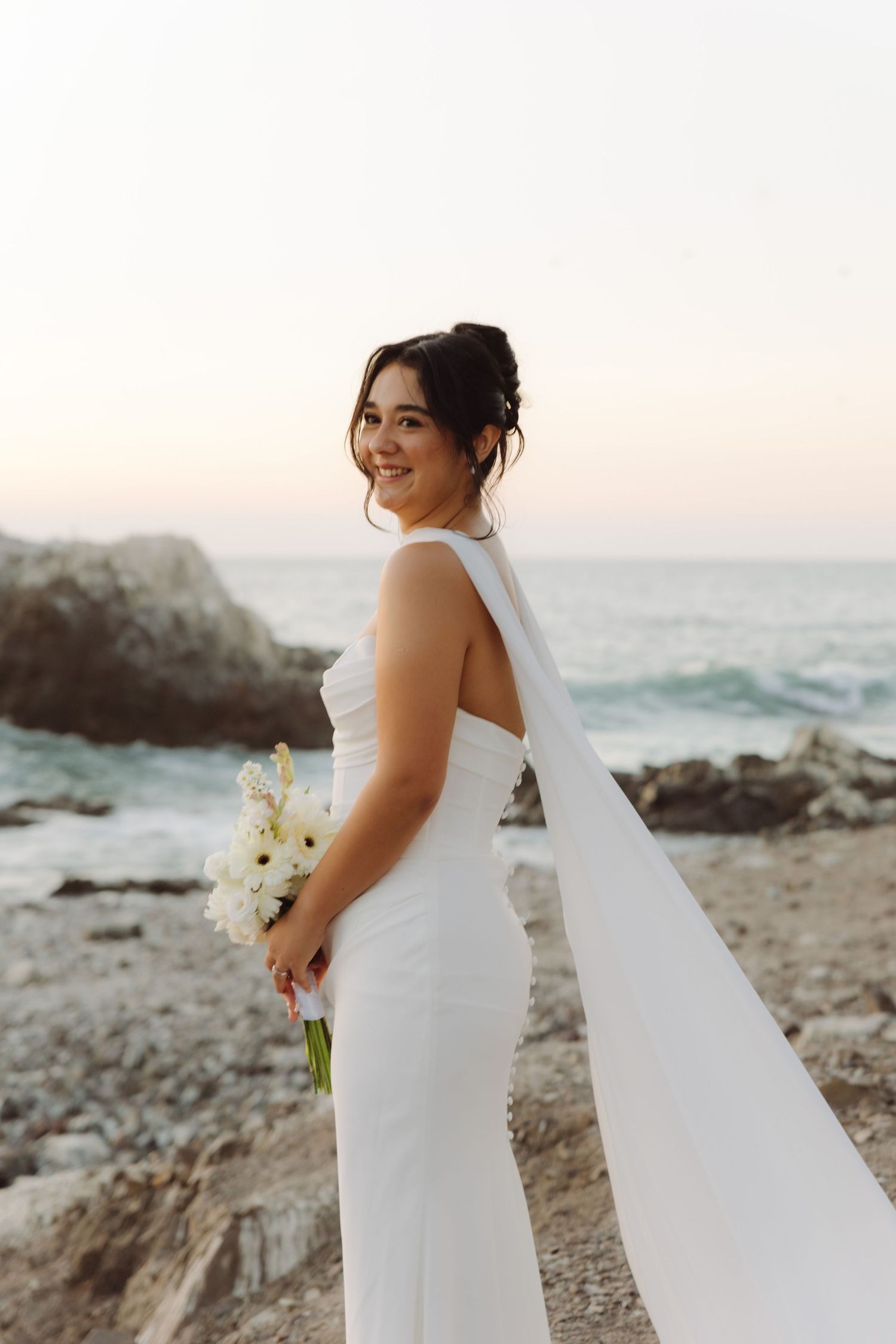 Smiling bride in a white halter wedding dress holds a white floral bouquet on a scenic rocky beach at sunset.