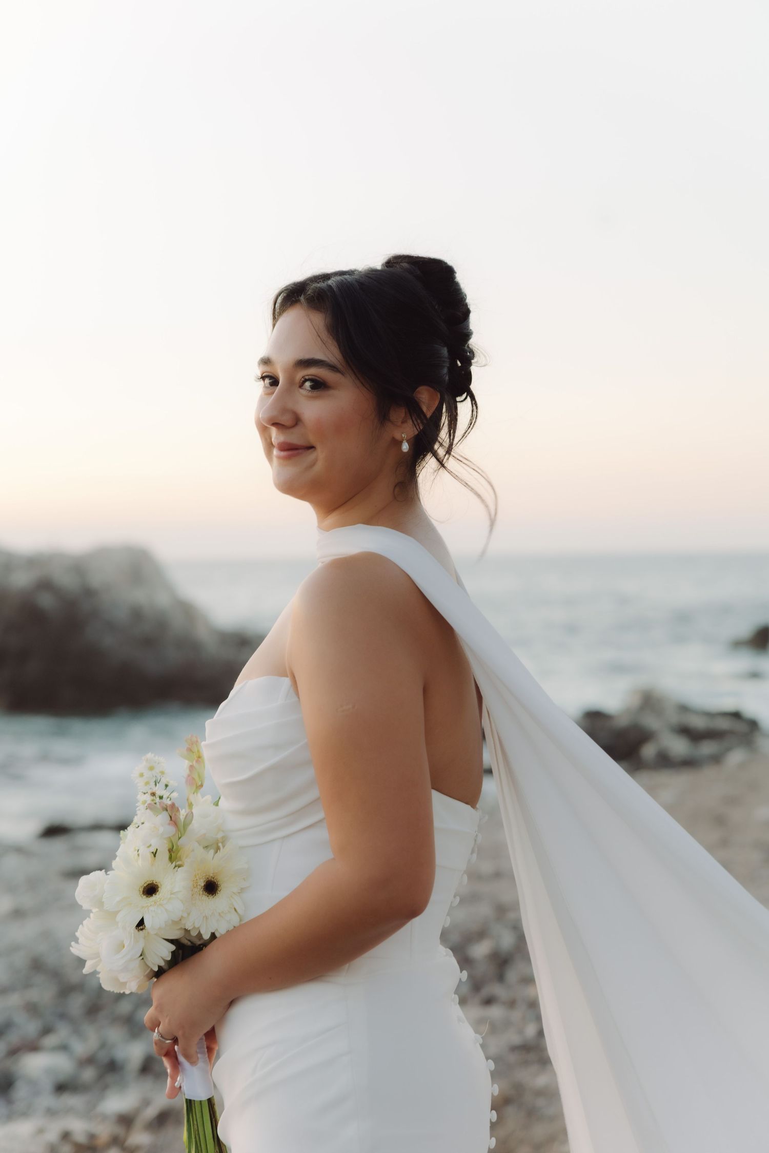 Beautiful bride in a white halter gown with flowing veil smiles while holding a white bouquet on a rocky beach.