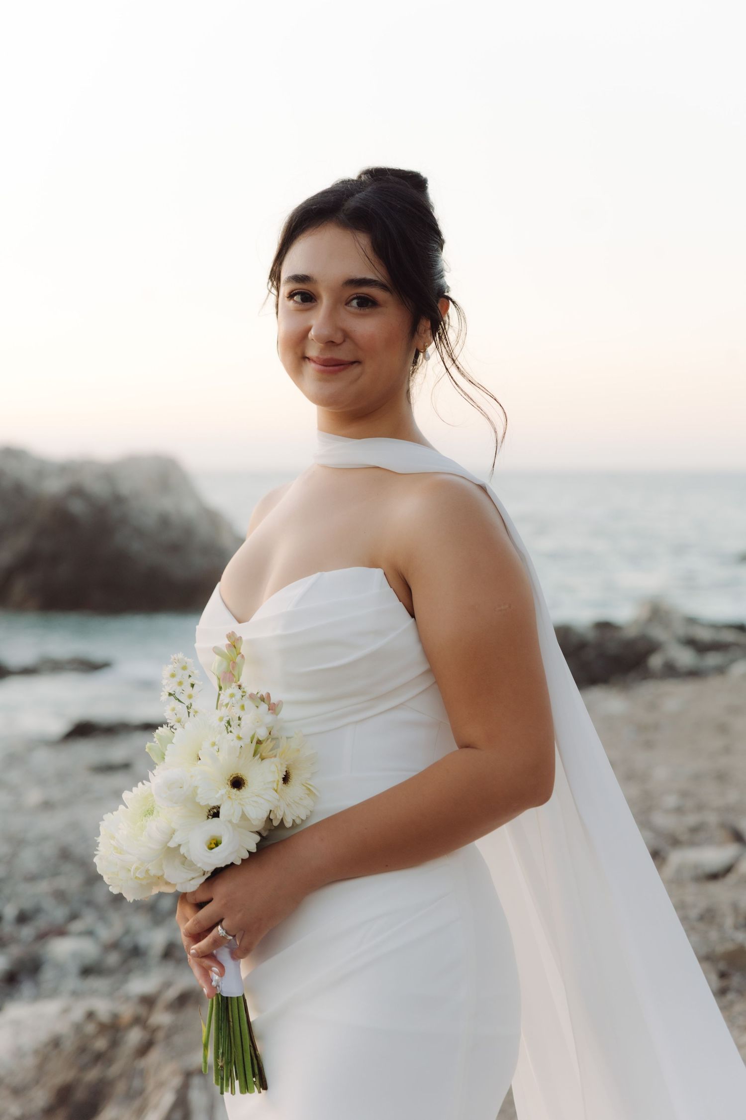Radiant bride in a white halter wedding dress holds a white gerbera daisy bouquet on a rocky beach at dusk.