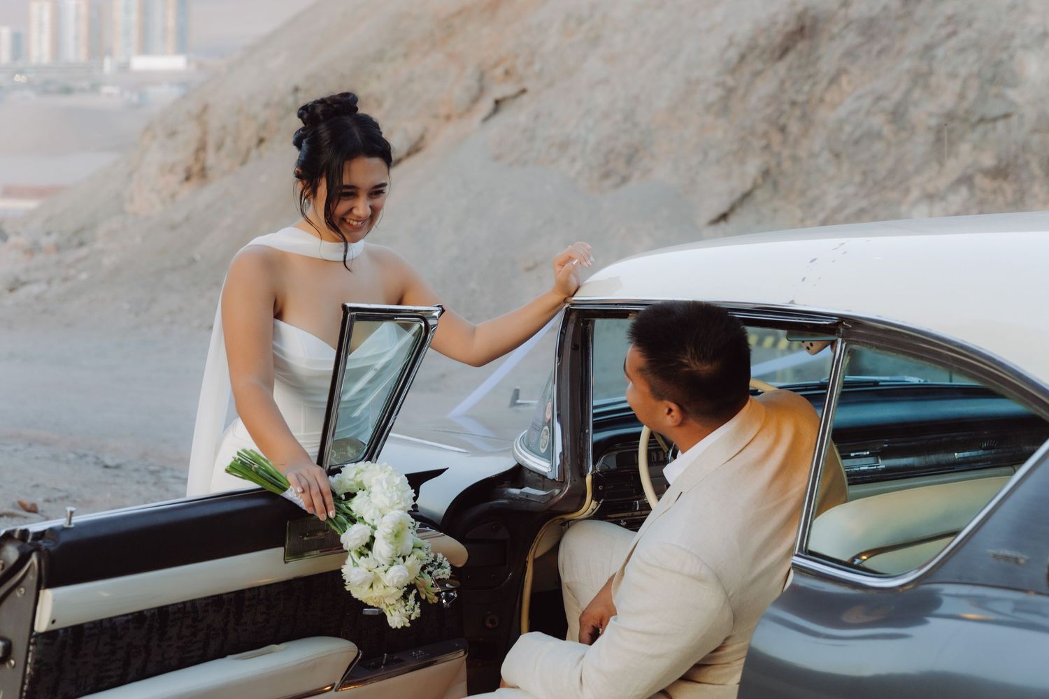 Smiling bride in a white strapless gown holds a bouquet while stepping out of a vintage car with the groom.