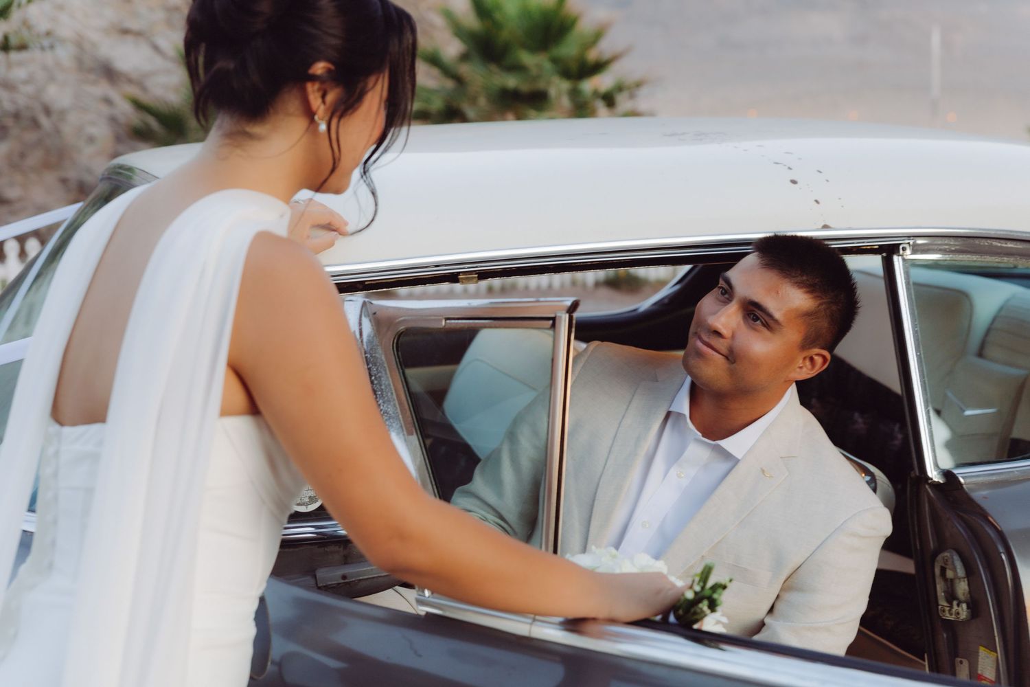 Groom in a beige suit smiles at the bride from inside a vintage car during their wedding day photo session.