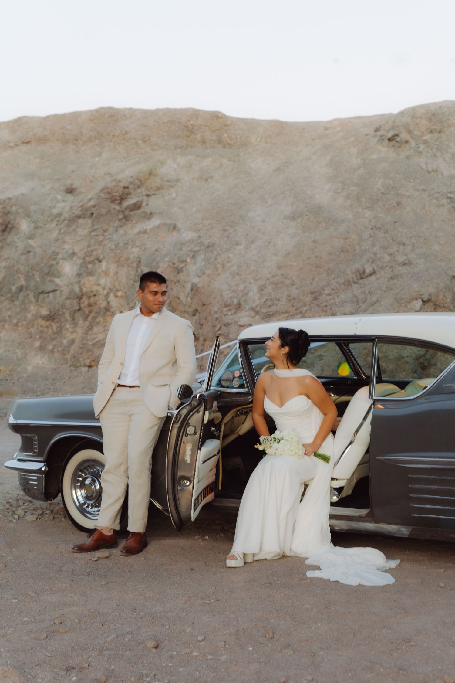 Bride and groom posing with a vintage classic car in a desert landscape during their wedding photo shoot.