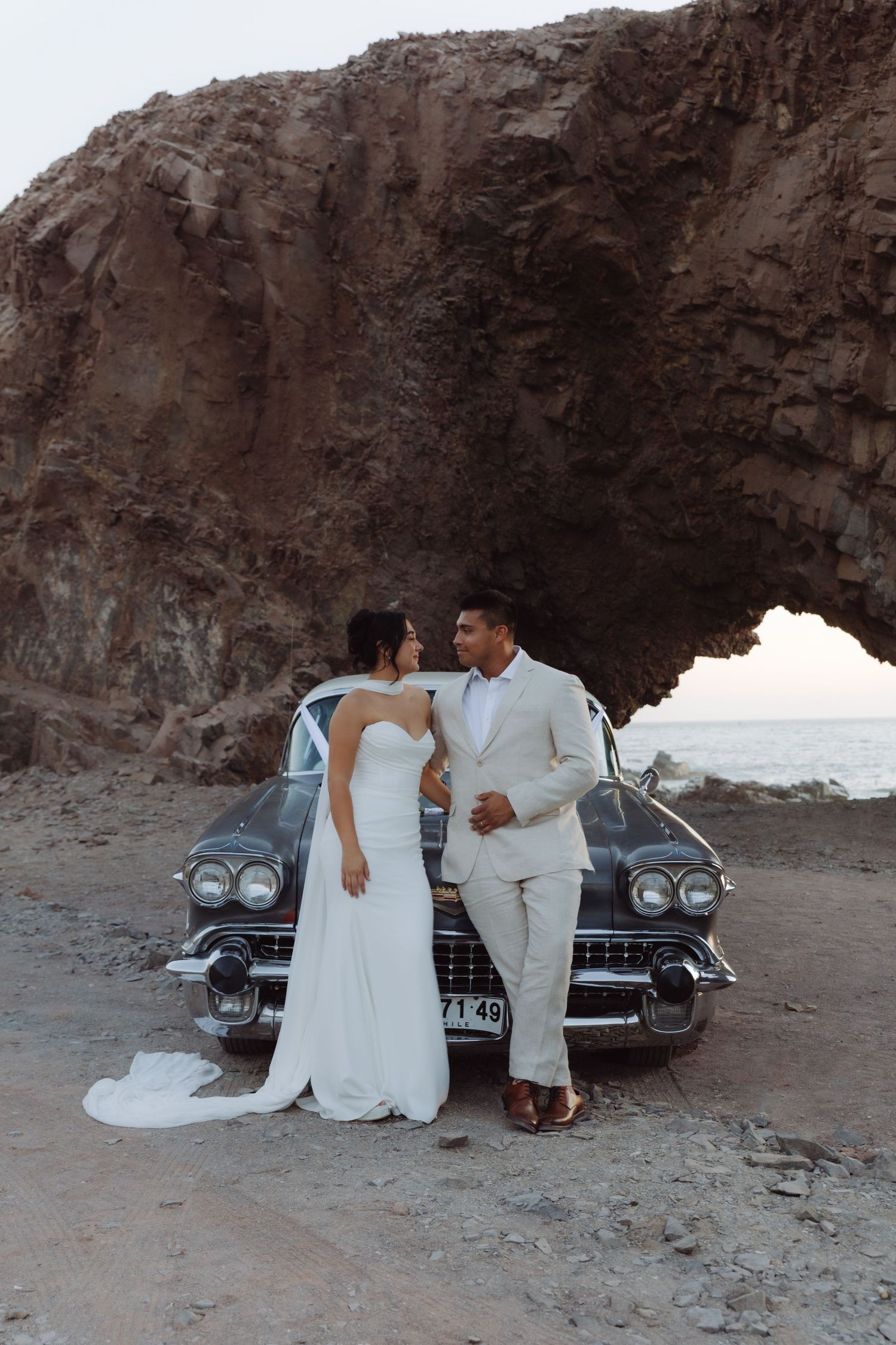 Wedding couple sharing a romantic moment leaning against a vintage Cadillac beneath a natural rock arch by the sea.
