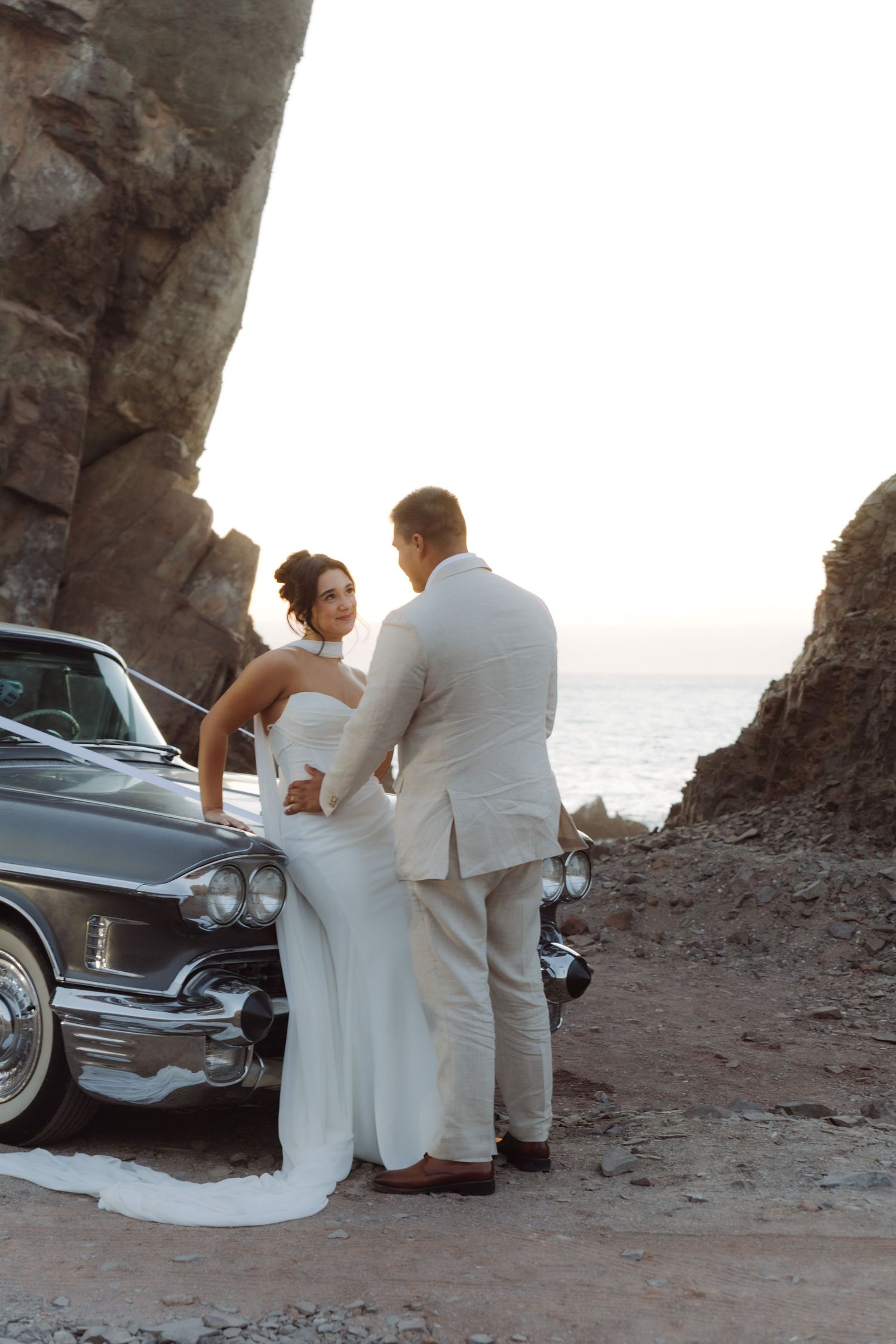 Bride and groom embracing beside a classic vintage car framed by dramatic coastal rock formations at sunset.