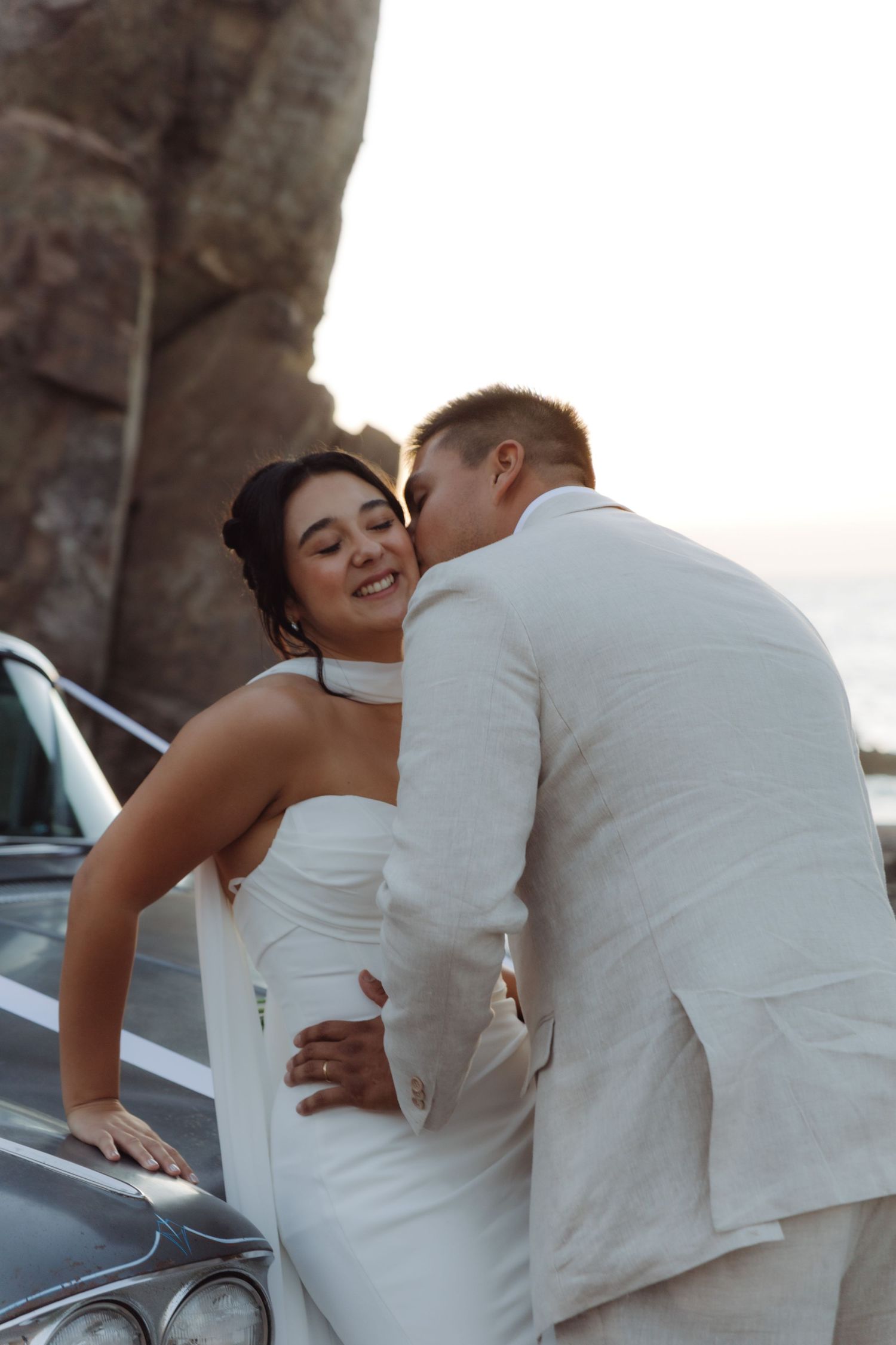 Groom kissing bride on the cheek as she smiles joyfully while leaning on a vintage car at golden hour.