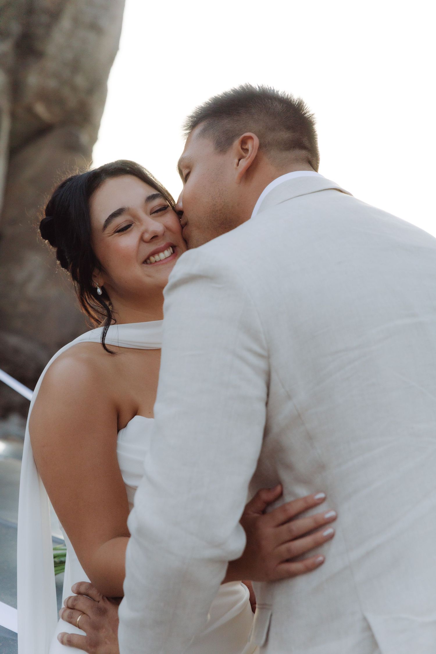 Groom tenderly kissing smiling bride on the cheek during romantic outdoor wedding photo session near rocky cliffs.
