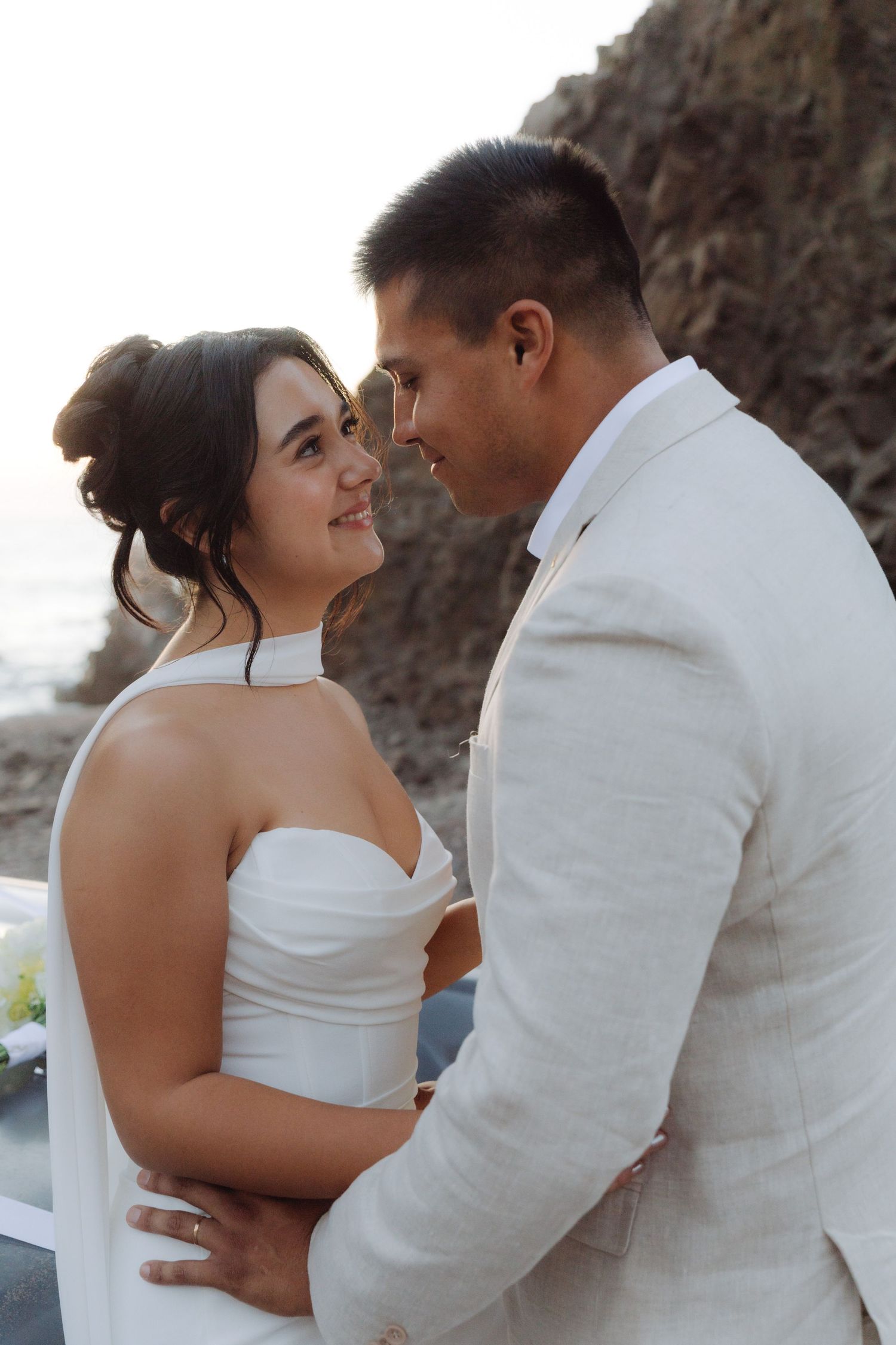 Bride and groom gazing into each other eyes while embracing during a sunset wedding photo session by the ocean.