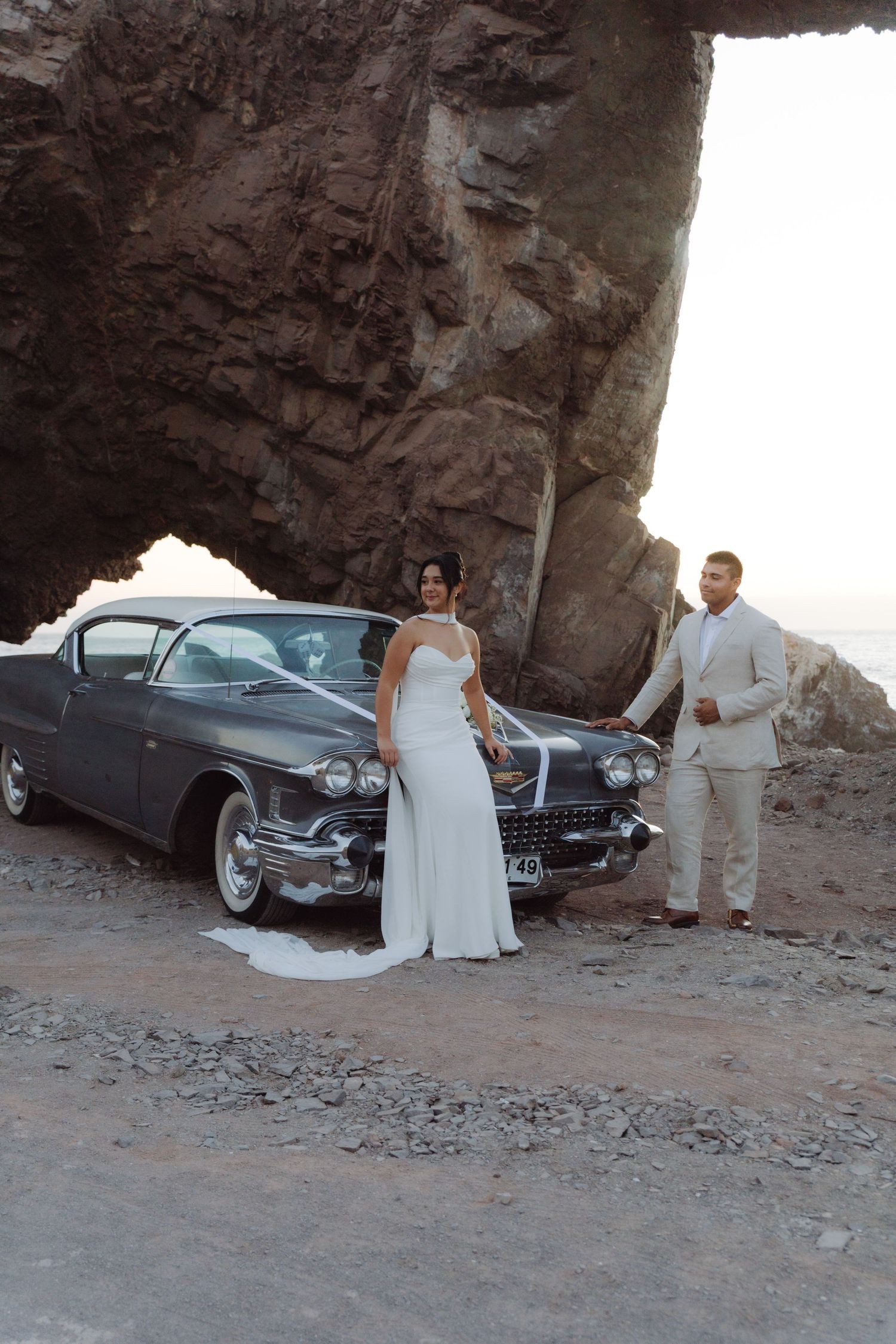 Elegant bride and groom posing with a vintage 1958 Cadillac under a natural rock arch at a coastal wedding shoot.