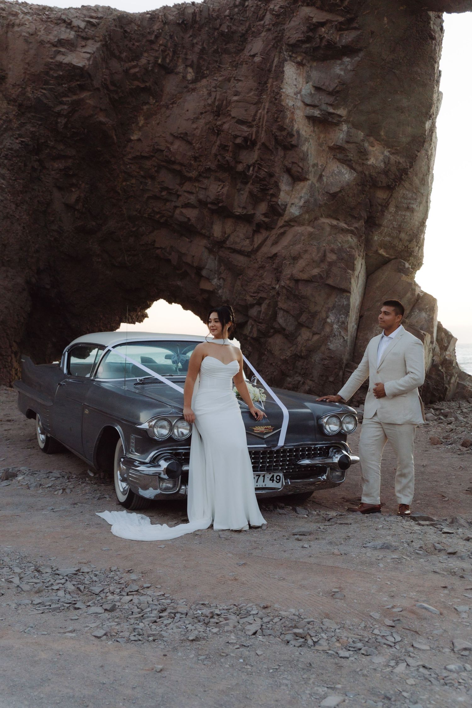 Wedding couple standing beside a decorated vintage Cadillac beneath a dramatic natural rock arch near the ocean.